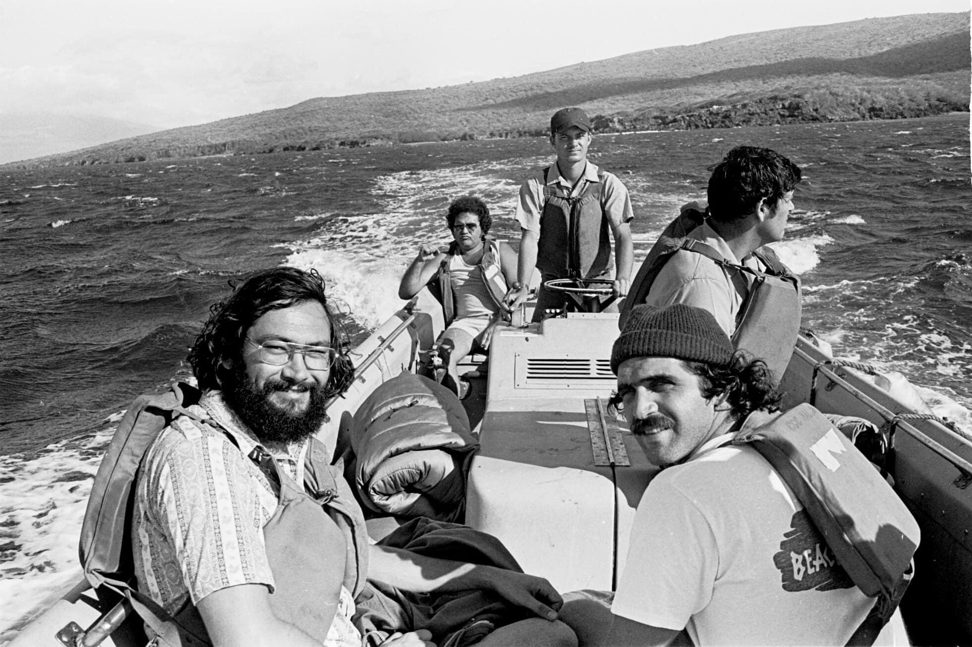 Seated in a boat, four male protestors in life vests are escorted from Kahoʻolawe’s Kūheʻeia Bay by a member of the U.S. Coast Guard.
