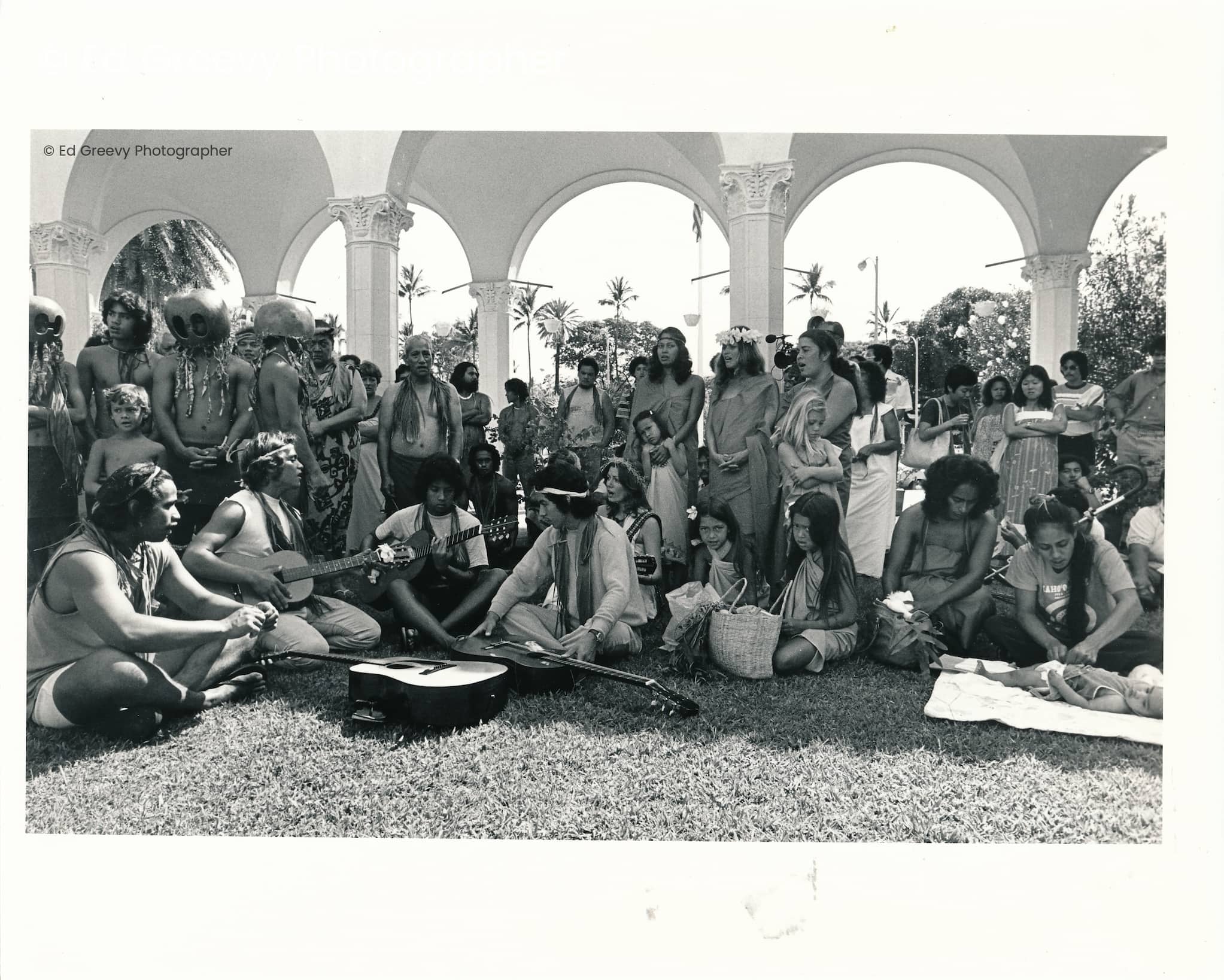 Members and supporters of Protect Kahoʻolawe ʻOhana gather on lawn outside of a federal courthouse with instruments. Some sing and some wear mākini.