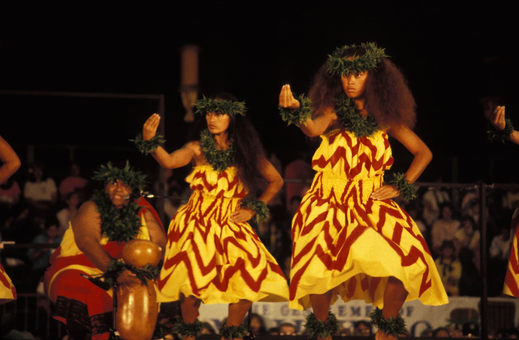 Two women dancers perform hula kahiko. Behind them, a male kumu hula is seated while singing into a microphone and playing ipu heke.