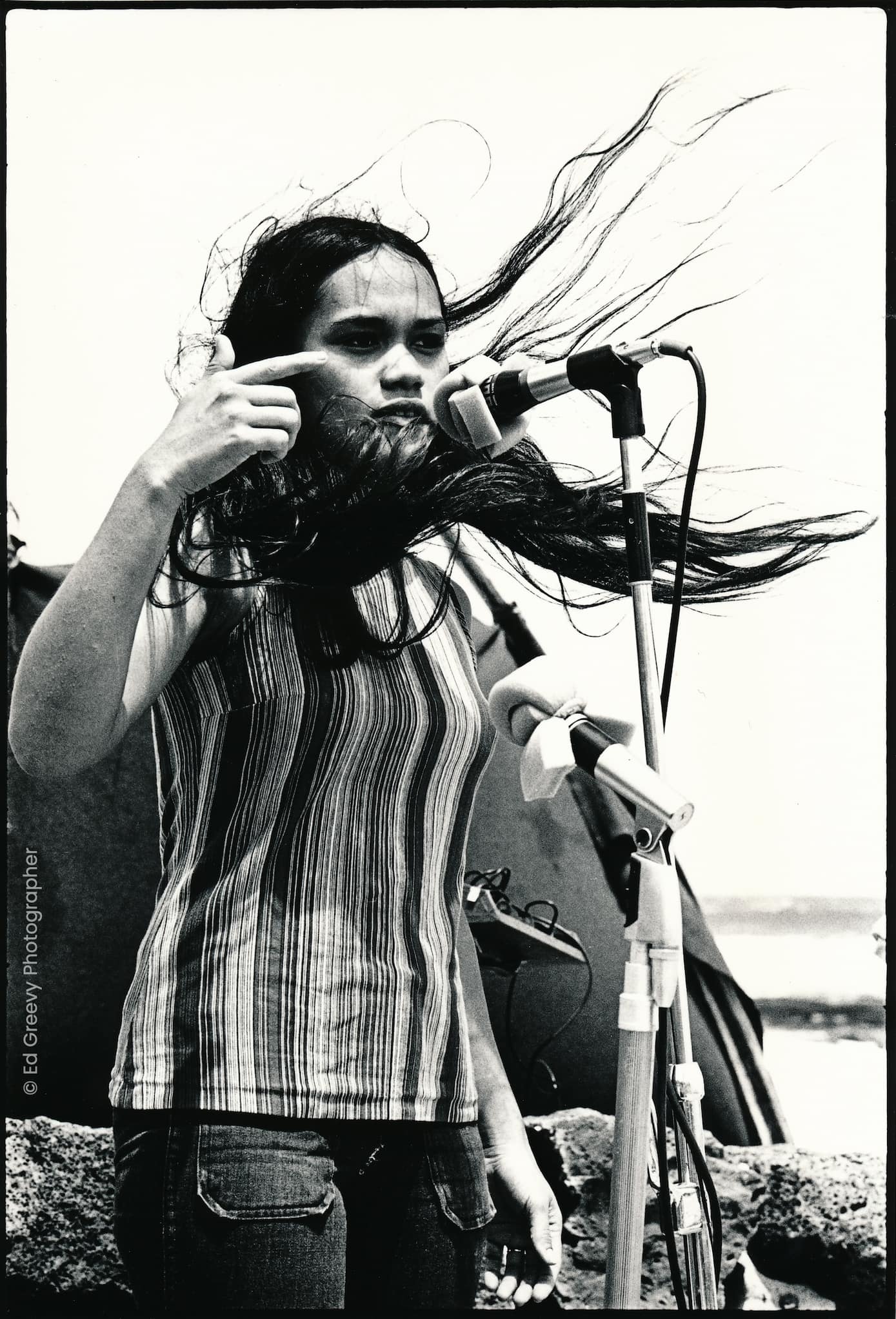 Kanaka ʻŌiwi activist Terrilee Kekoʻolani-Raymond speaks into a microphone during outdoor rally. Next to her is a white flag embroidered with "SOS."