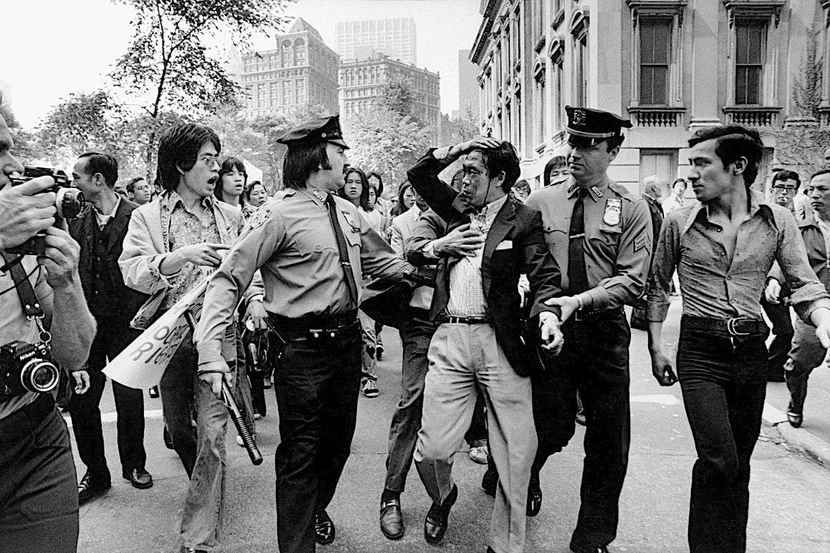 Police officers stand on either side of protestor bleeding from head wound. The police officers and protestor are surrounded by crowd of onlookers.