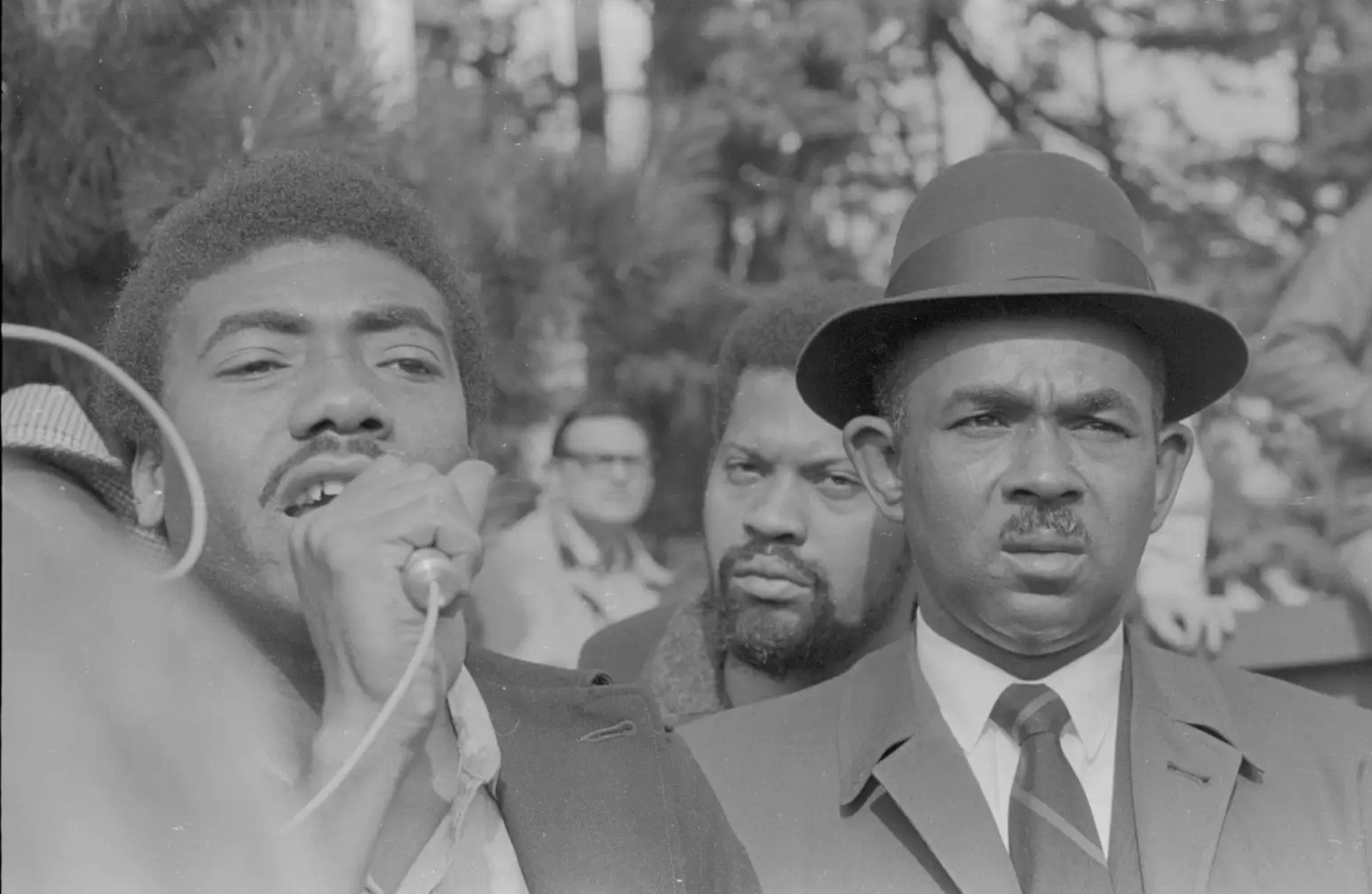 A trio of serious-looking black men stand outside among onlookers. One man speaks into a microphone.