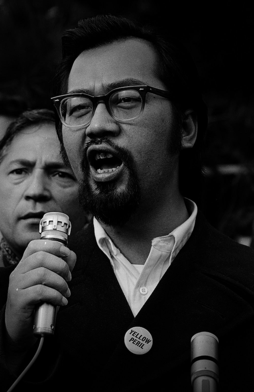 George Woo shouts into a microphone. He wears a button with the slogan yellow peril.