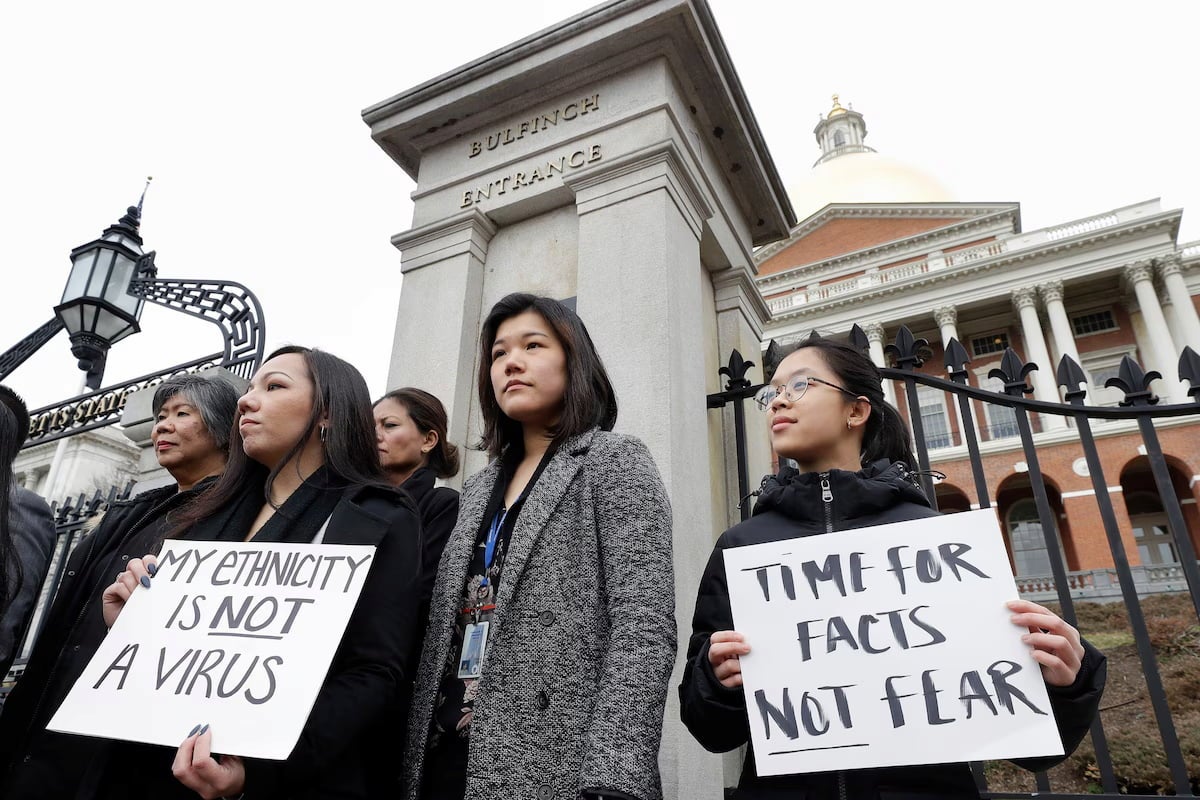 A group of Asian women stand at the Bulfinch Entrance holding signs protesting the association of ethnicity with a virus.