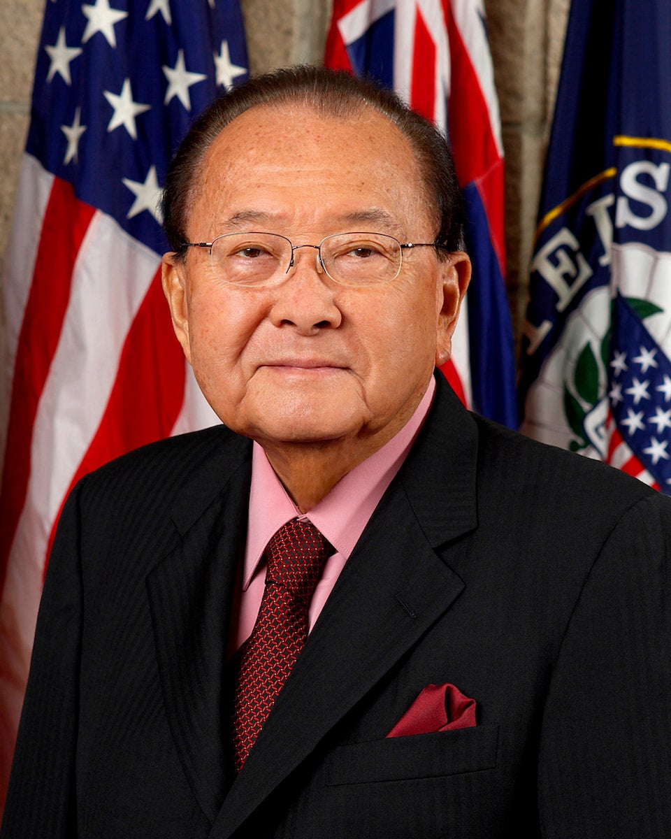 Formal portrait of Daniel Inouye with flags as a background.