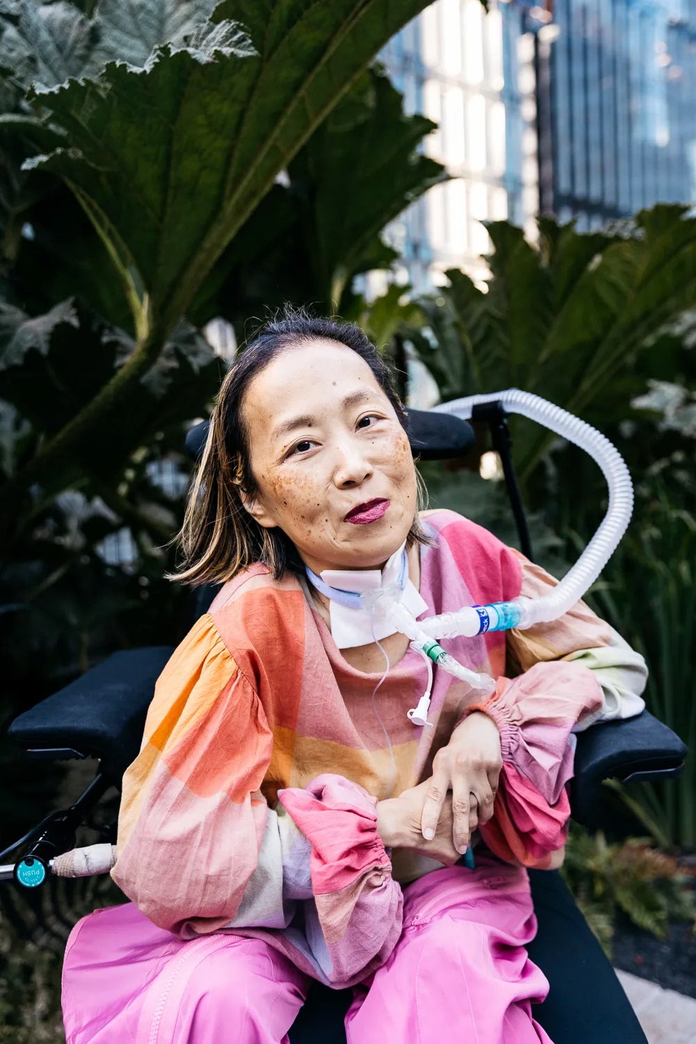 Alice Wong sits outside with a lush background and wears a colorful outfit. Her trach extends from her throat to the back of the wheelchair.