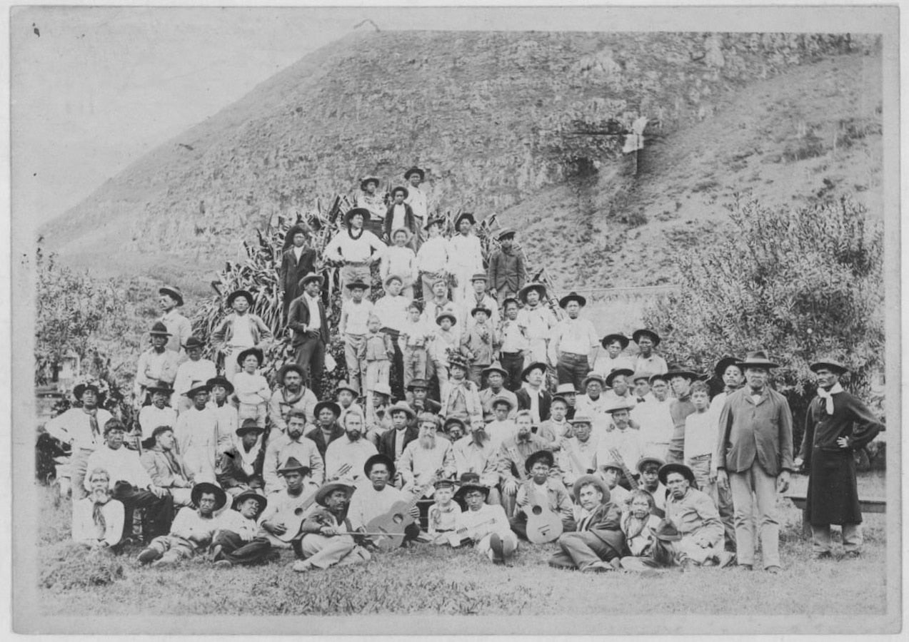 A group of men pose on a barren mountain side. Most wear wide brimmed hats and light-colored shirts.