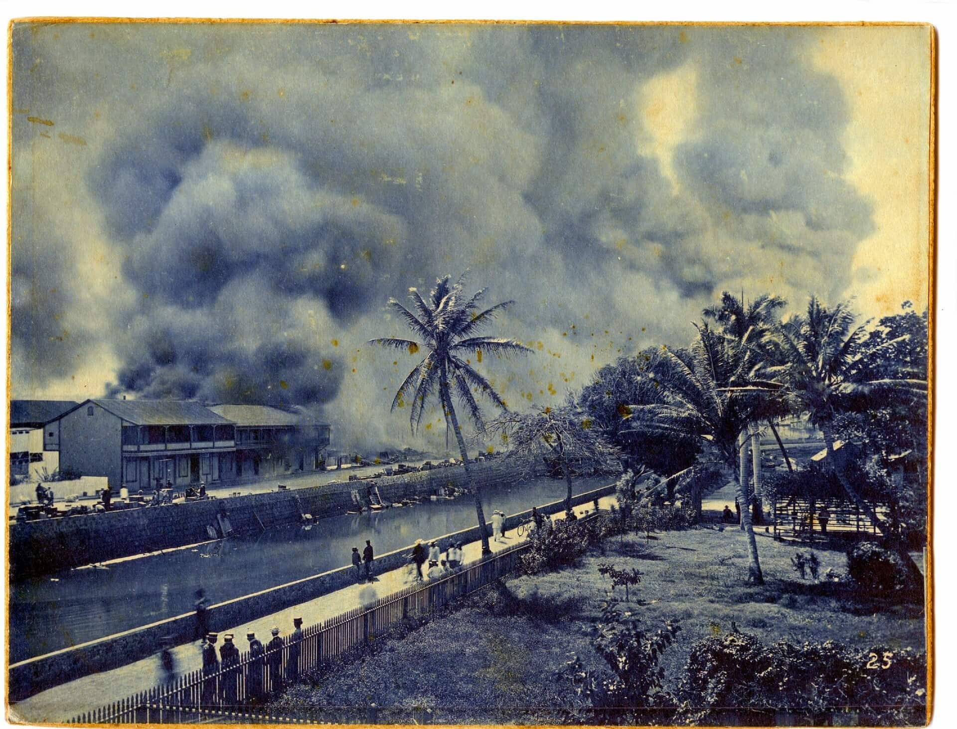 A river divides a scene. One side is obliterated with smoke. The other has pedestrians strolling down a walkway lined with grass and palm trees.