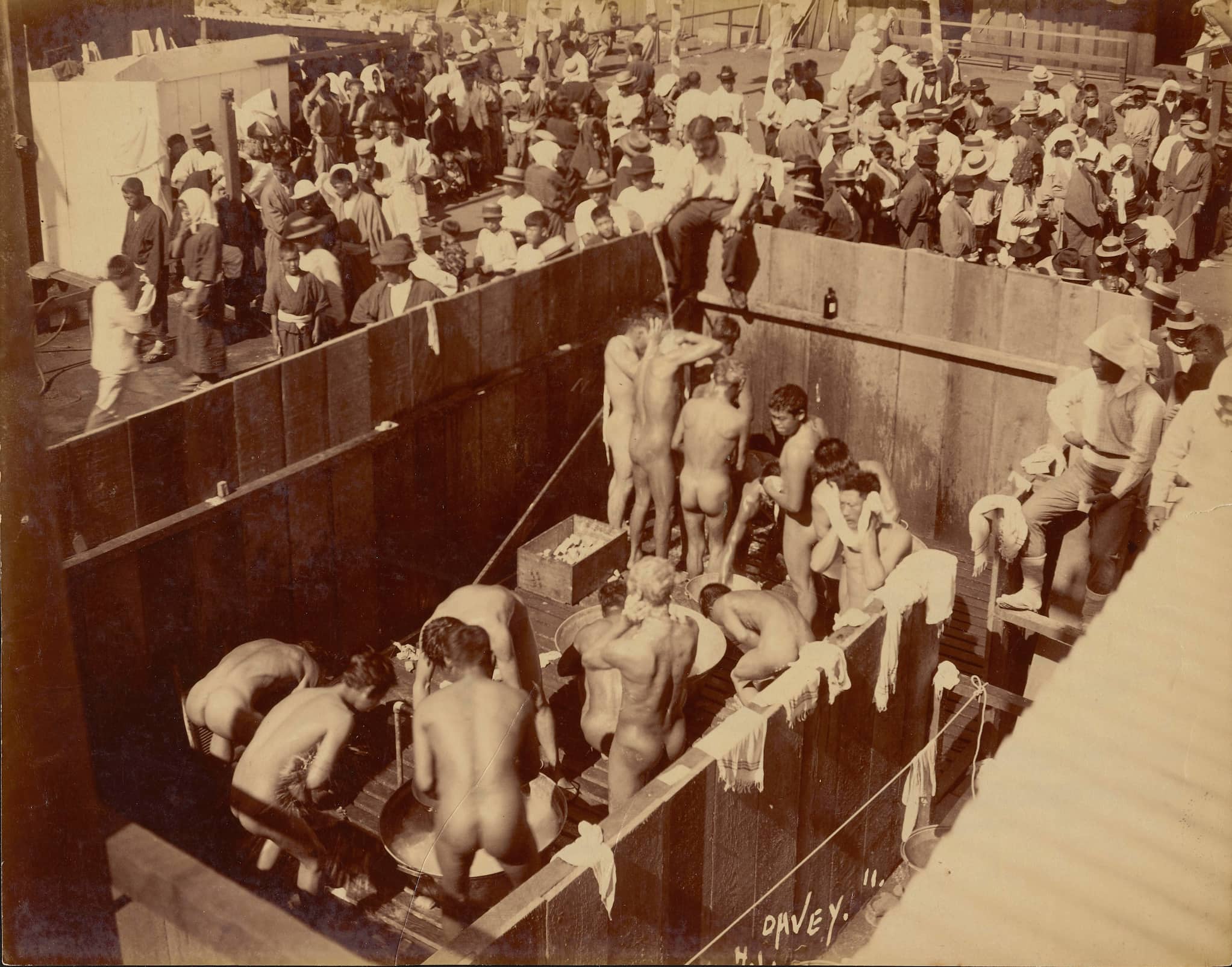 A large crowd of clothed people wait as naked men bathe en masse in a sunken area.