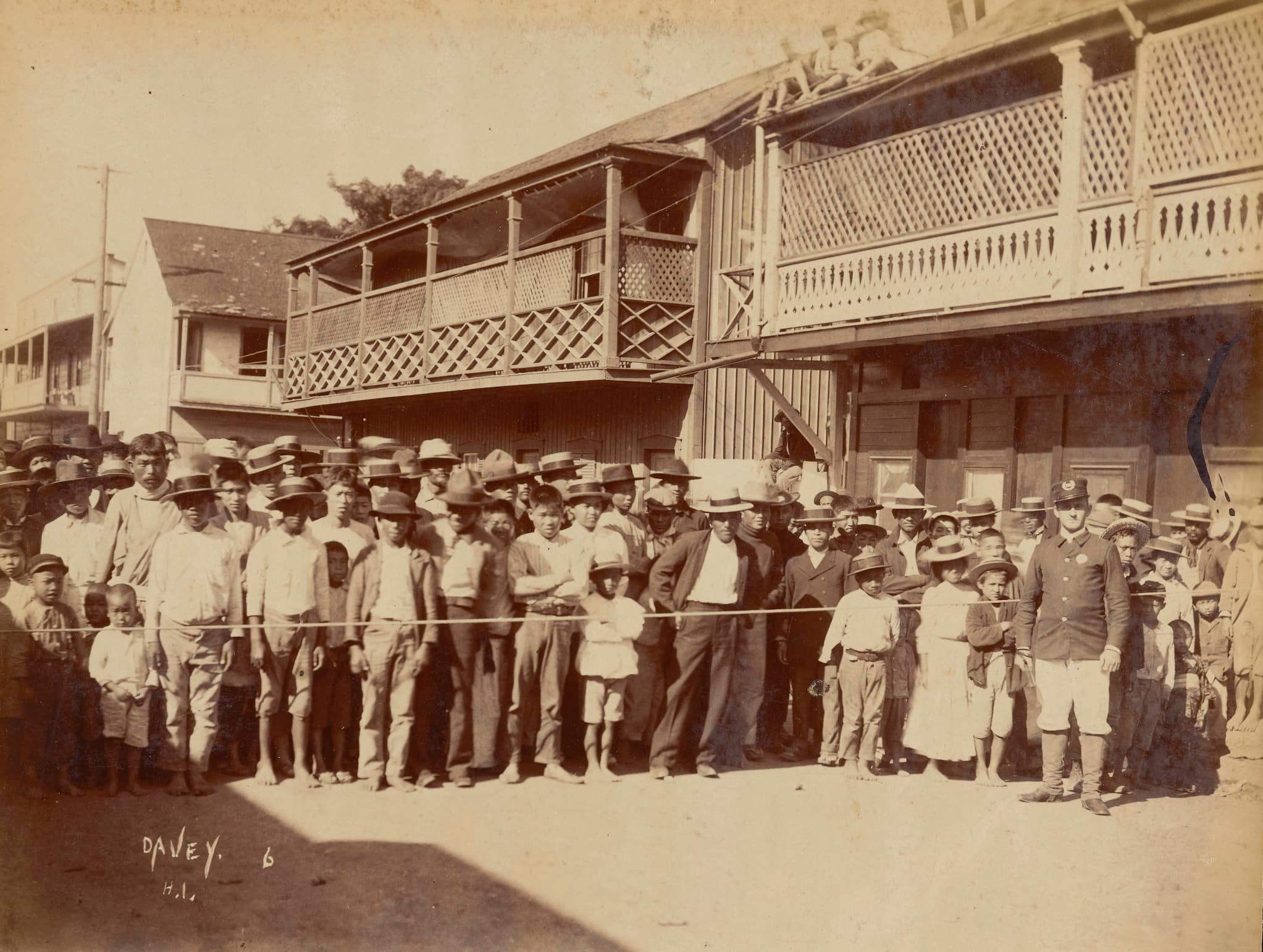 An officer stands in front of a rope. Crowds of families are packed behind the rope. The ramshackle housing forms the background.