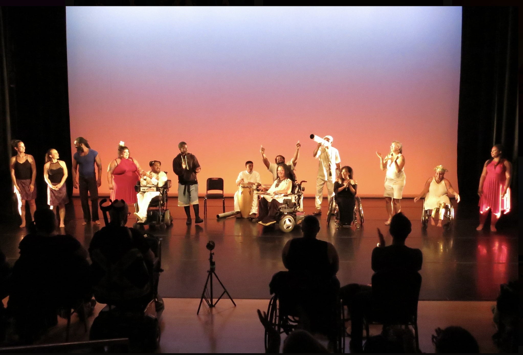 Actors line an orange-lit stage with no backdrop. Some actors and audience members are in wheelchairs.