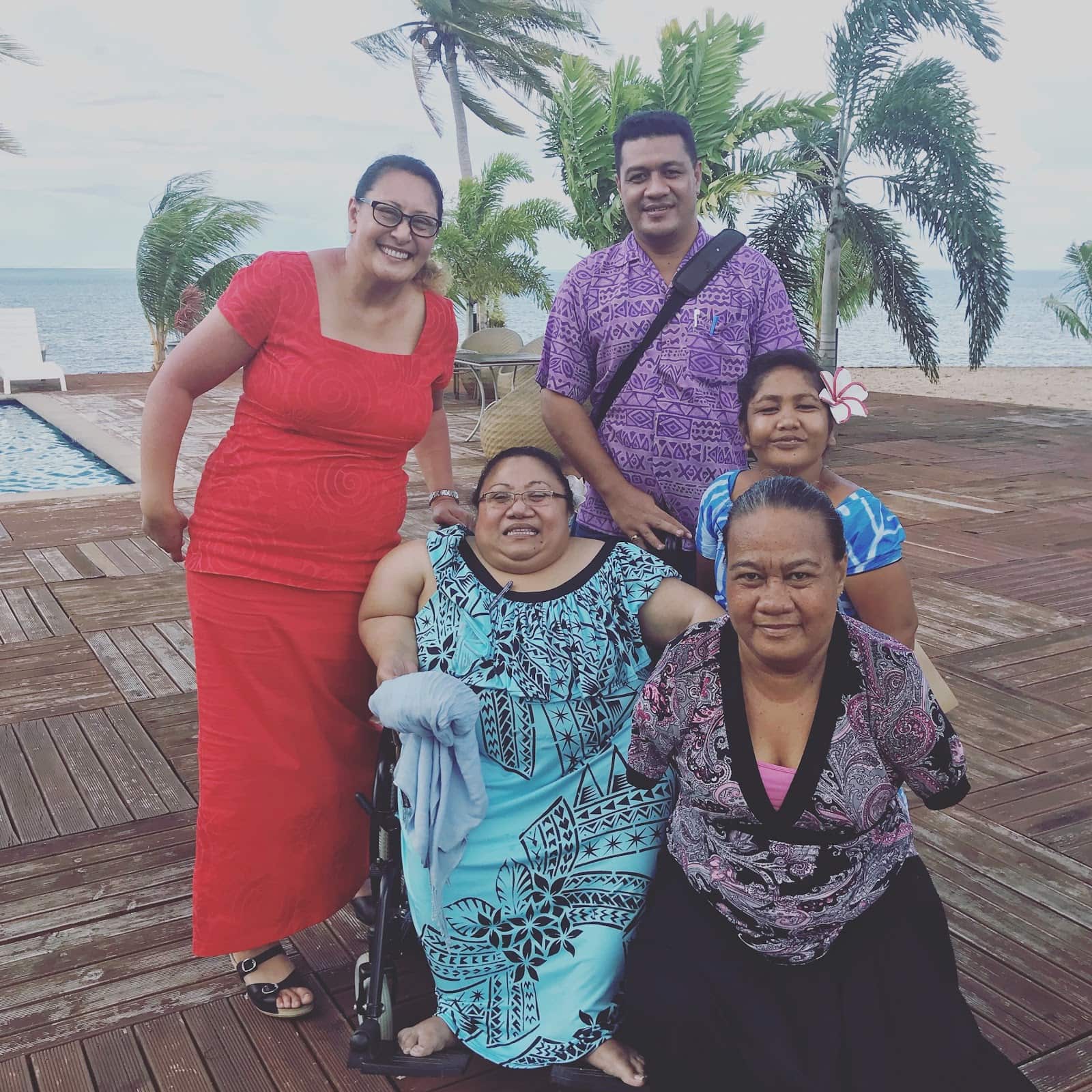A group of people pose with a woman seated in a wheelchair in the center and an armless woman in front. The ocean and palm trees are behind them.