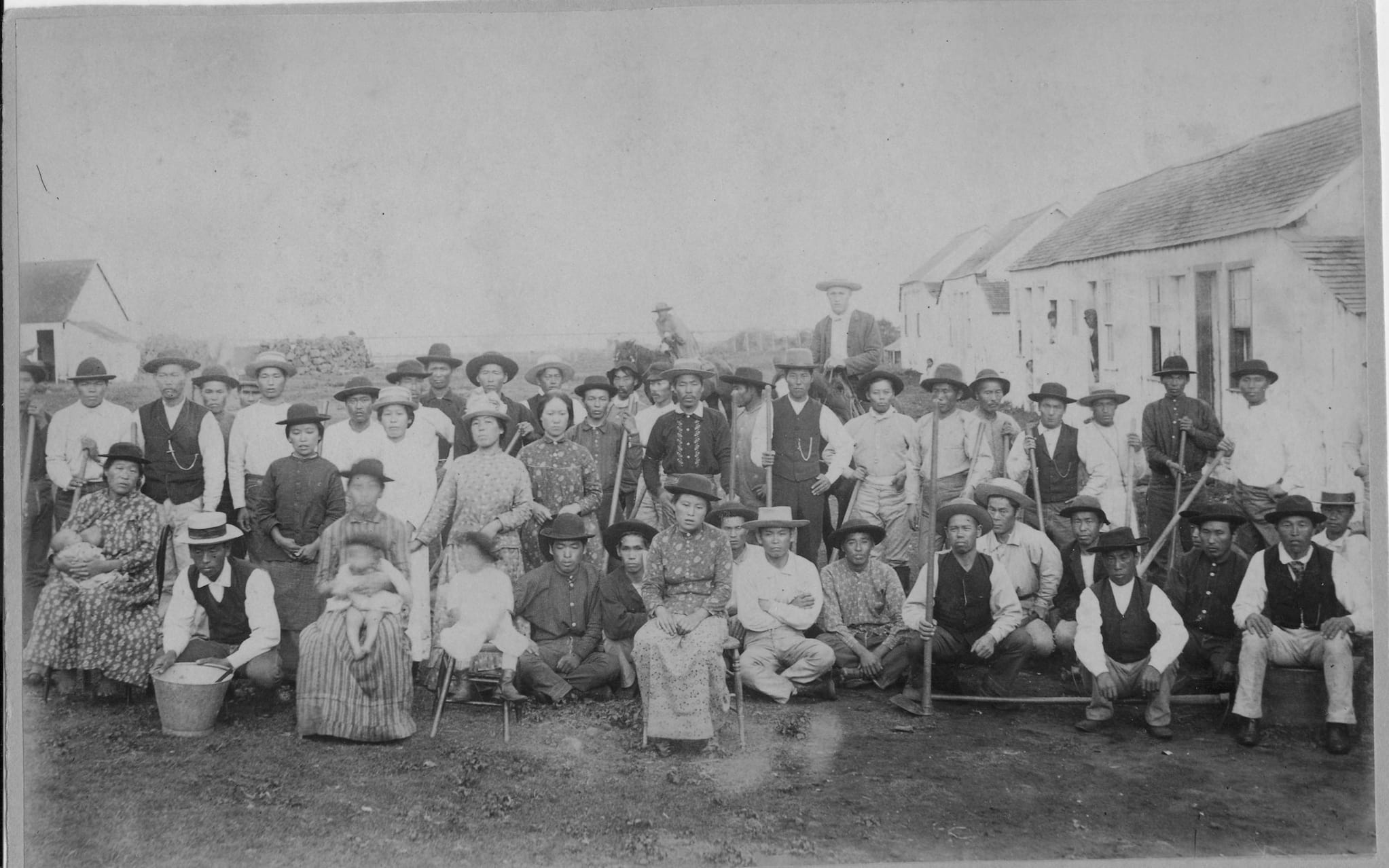 Plantation workers pose with their wives, children, and tools in front of group housing.