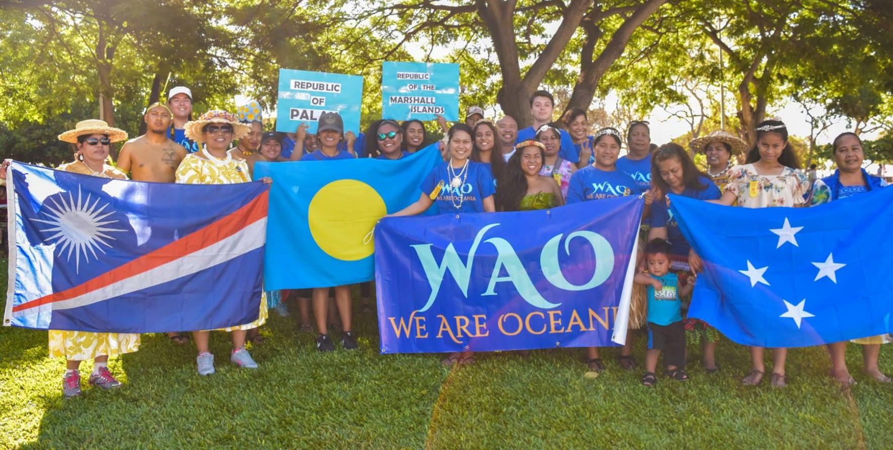 A group of people gather under the shade of the trees, smiling, and holding blue colored flags and signs in support of We Are Ocean.