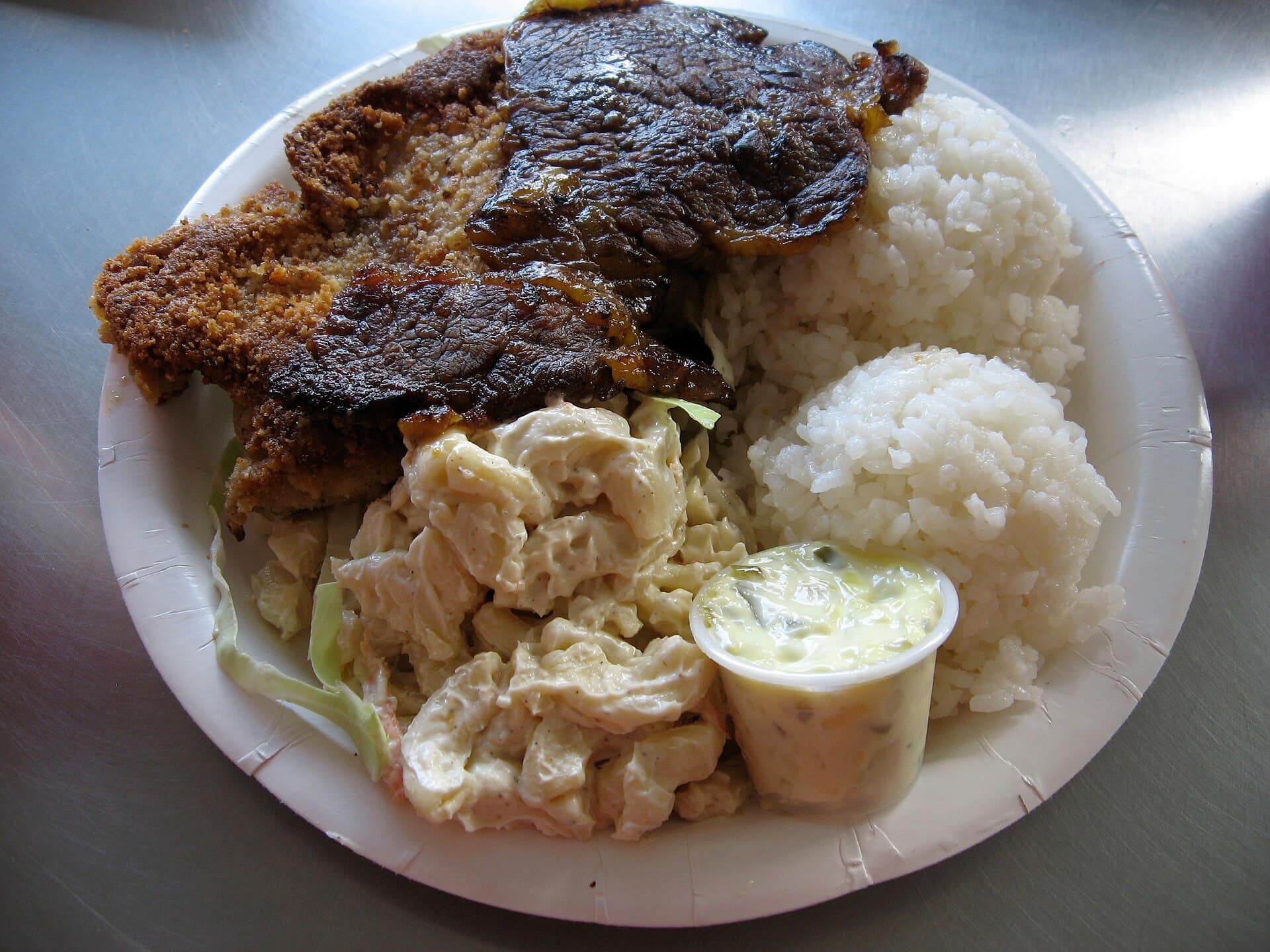 A plate stacked with mouth-watering pork, chicken and rice over some greens and served with a small cup of creamy macaroni salad.