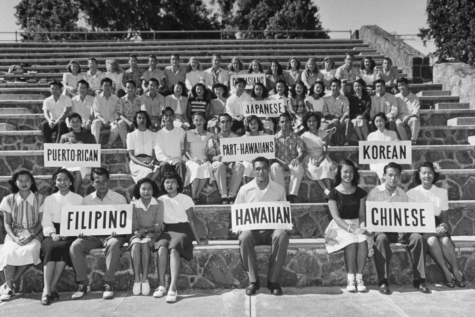 Students pose in groups on stone bleachers holding signs representing Filipino, Hawaiian, Chinese, Puerto Rican, Part-Hawaiians, Korean, Japanese, and Caucasians.