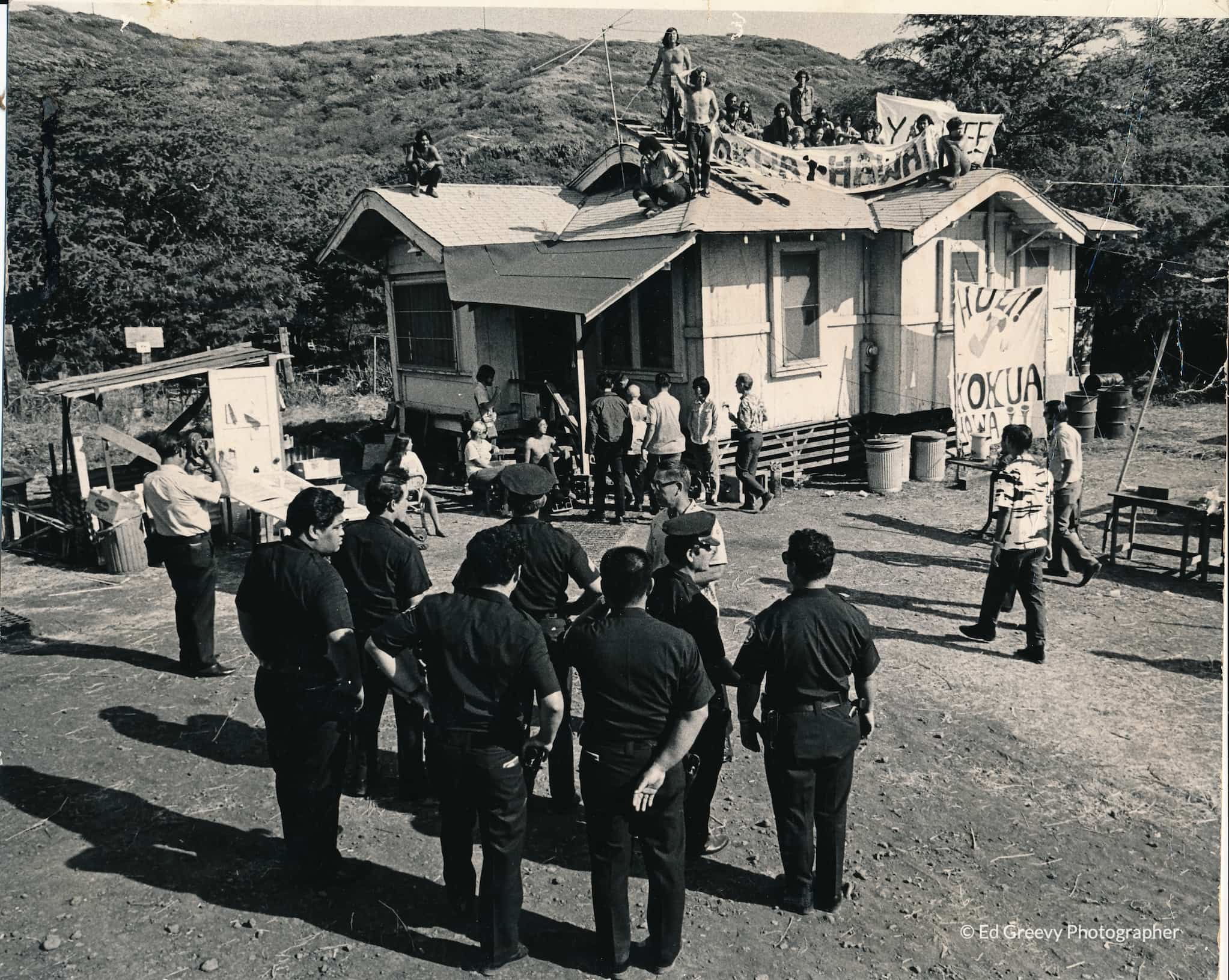 A group of police approach protesters sitting on the roof of a house with their banners.