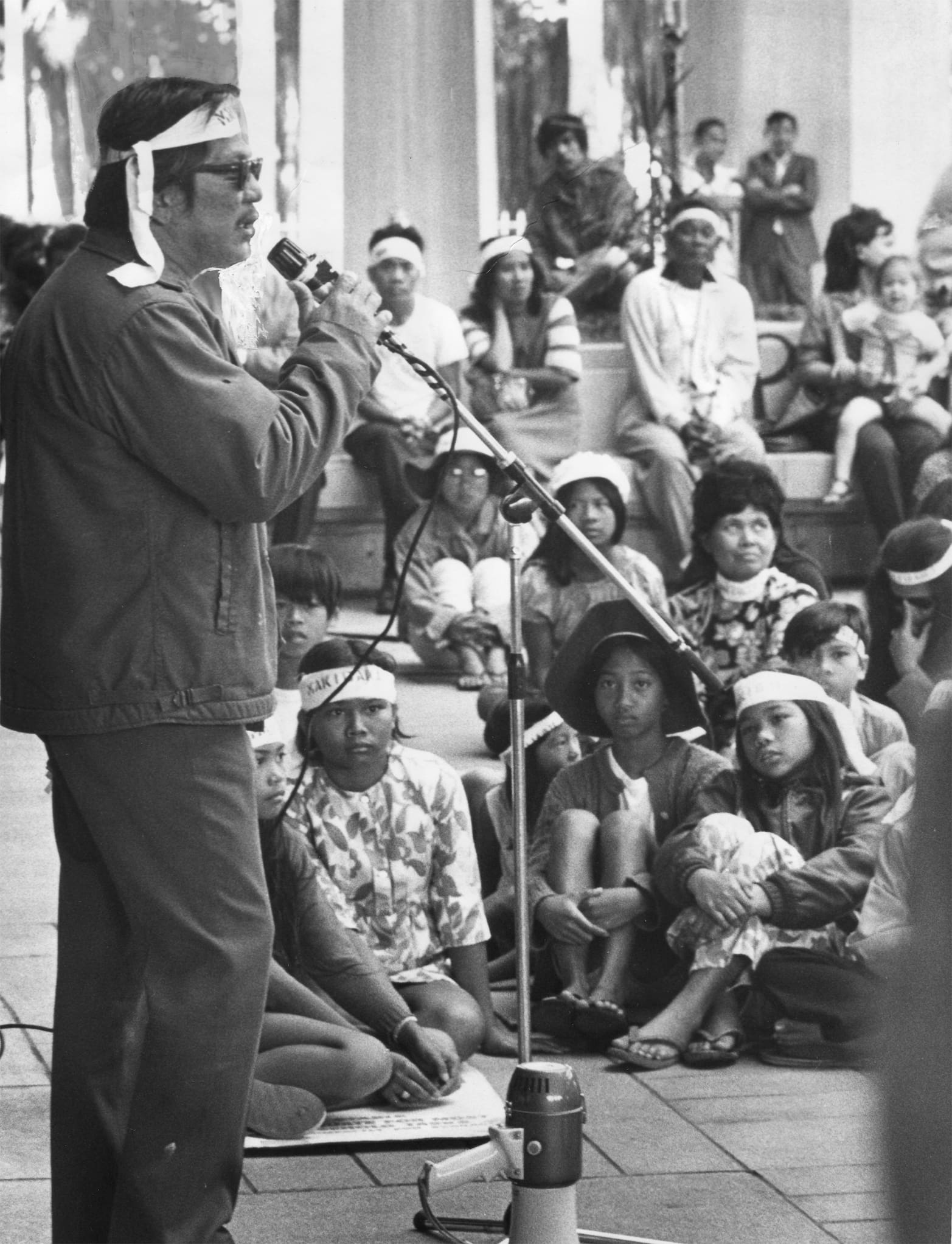 Pete Tagalog speaks to a seated crowd with children in the front. A megaphone rests next to the microphone stand.