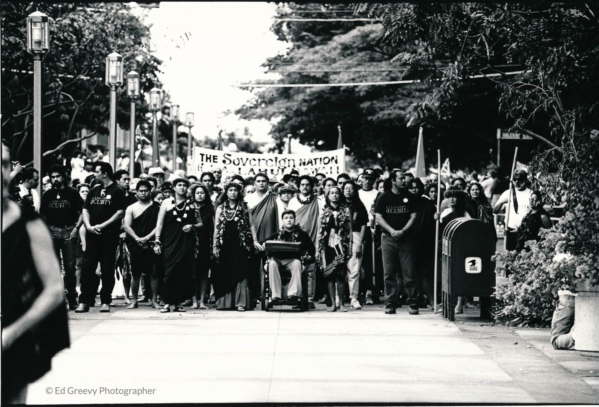 A march led by people in traditional dress and a young man in a wheelchair. The protesters span the wide walkway and are flanked by security.