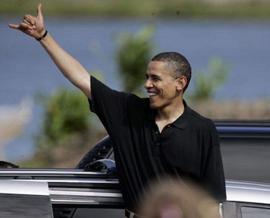 A casually dressed Barack Obama flashes the hand sign with his thumb and pinky up.