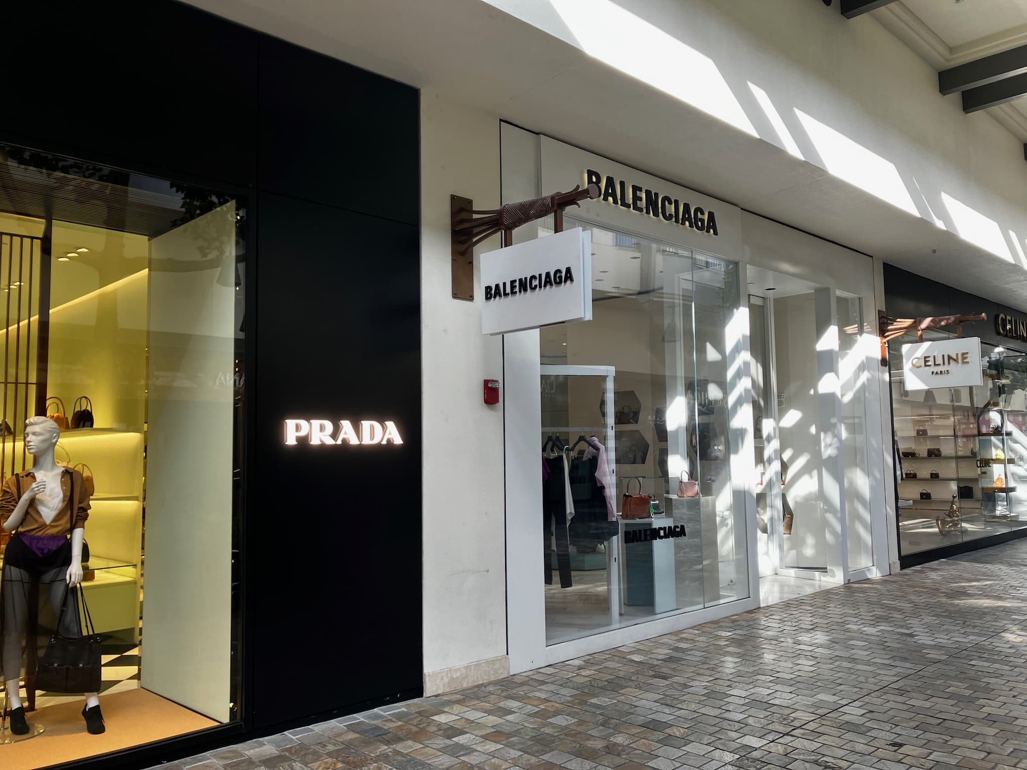 A row of upscale store fronts displaying signs for Prada, Balenciaga, and Celine.