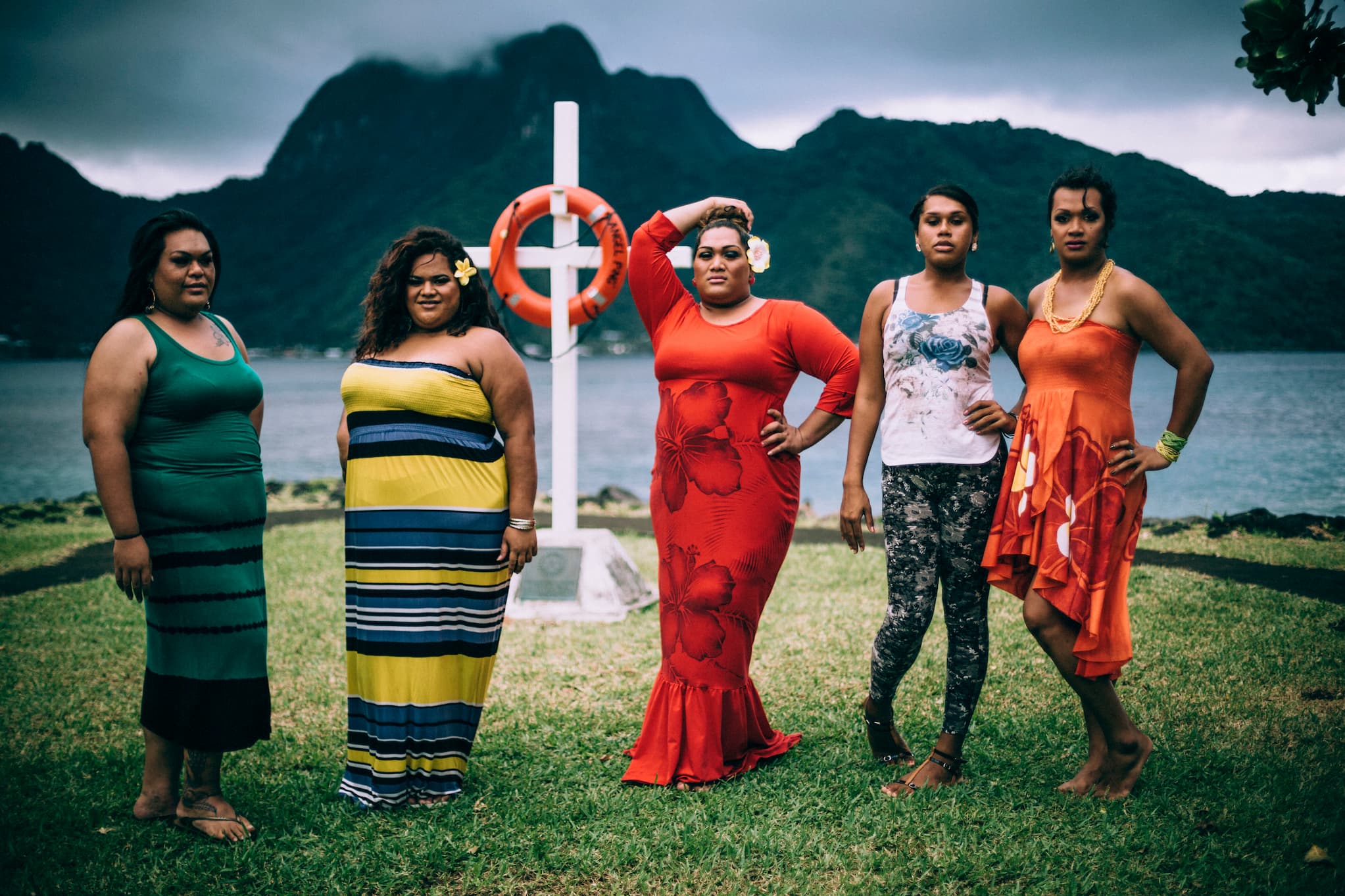A group of Fa'afafine pose together outside in front of a cross with a wreath. The background is a mountain across from the water.