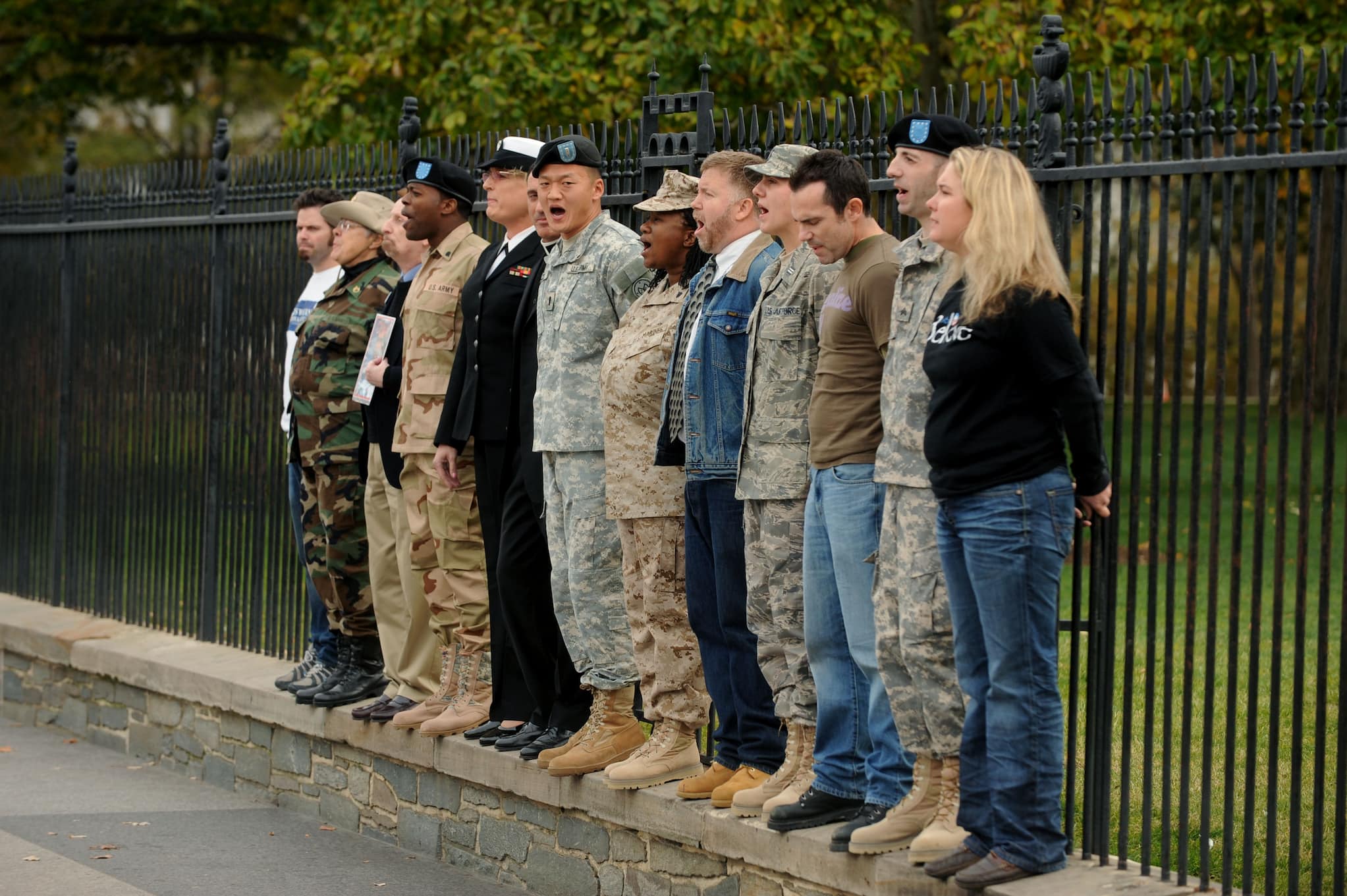 Civilians and military members in different uniforms stand shoulder to shoulder against a black post fence.