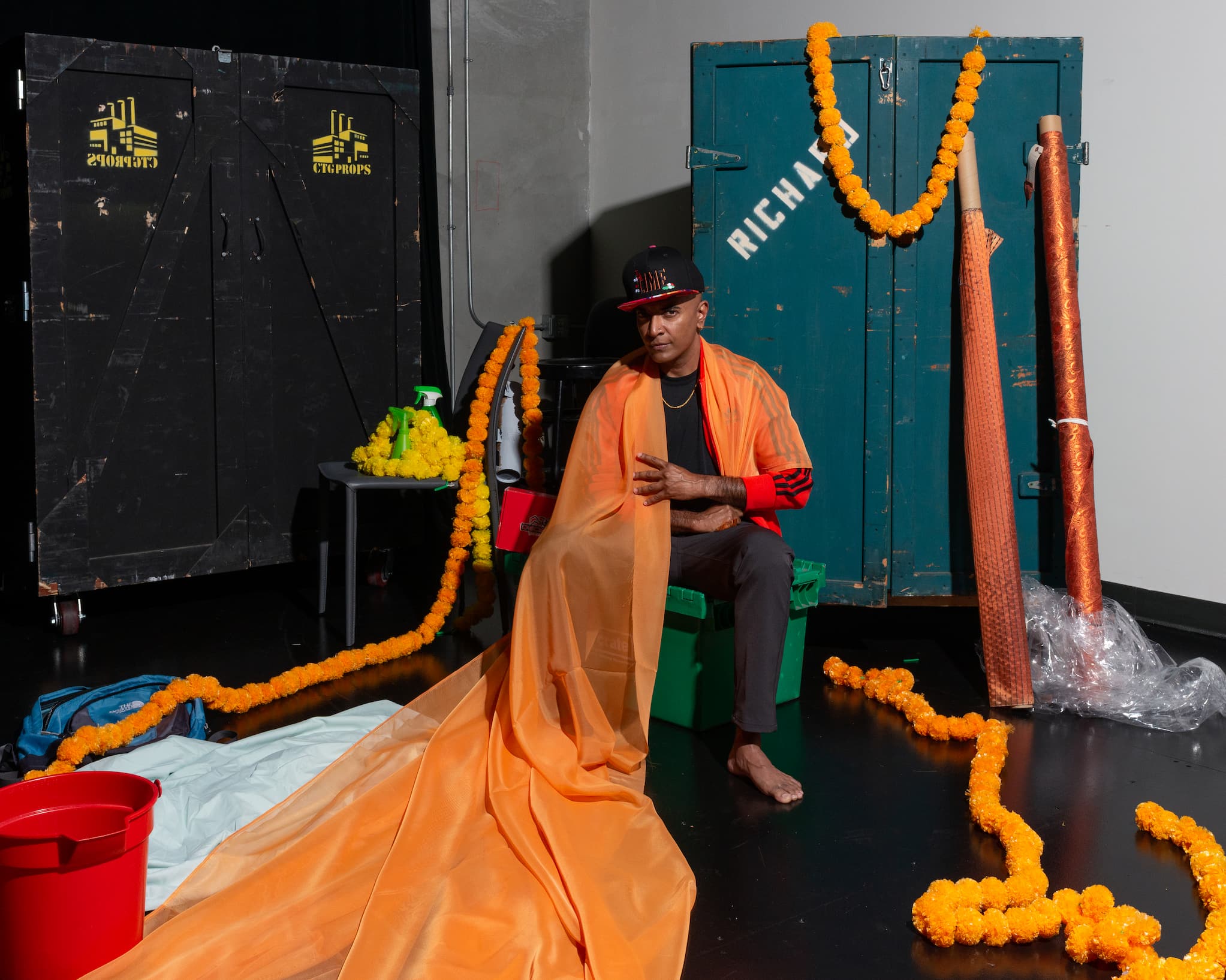 D’Lo sits on a bin surrounded by old lockers, spools of shiny orange fabric and orange flower garlands. He is draped in a long piece of orange fabric.