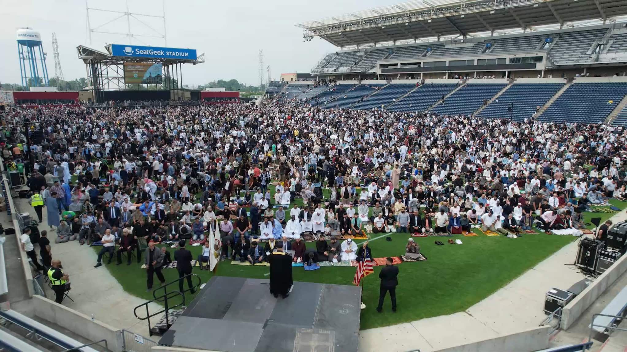 A large crowd of people, gathered across grass field at stadium, face towards a man standing on a small stage set up in the foreground.