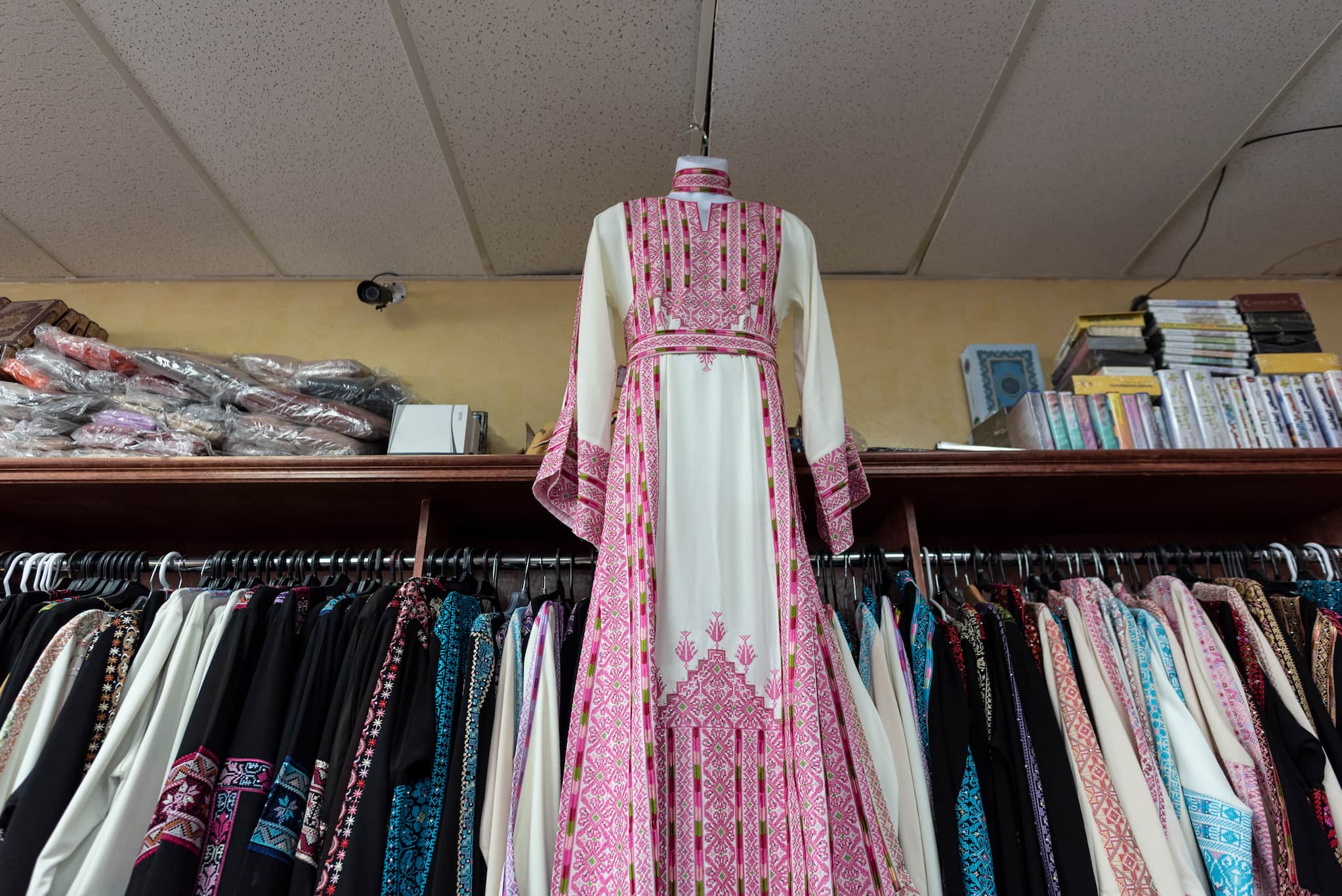 A white, long-sleeved dress with intricate pink and green embroidery, is suspended by a hanger above a clothing rack with similar dresses.