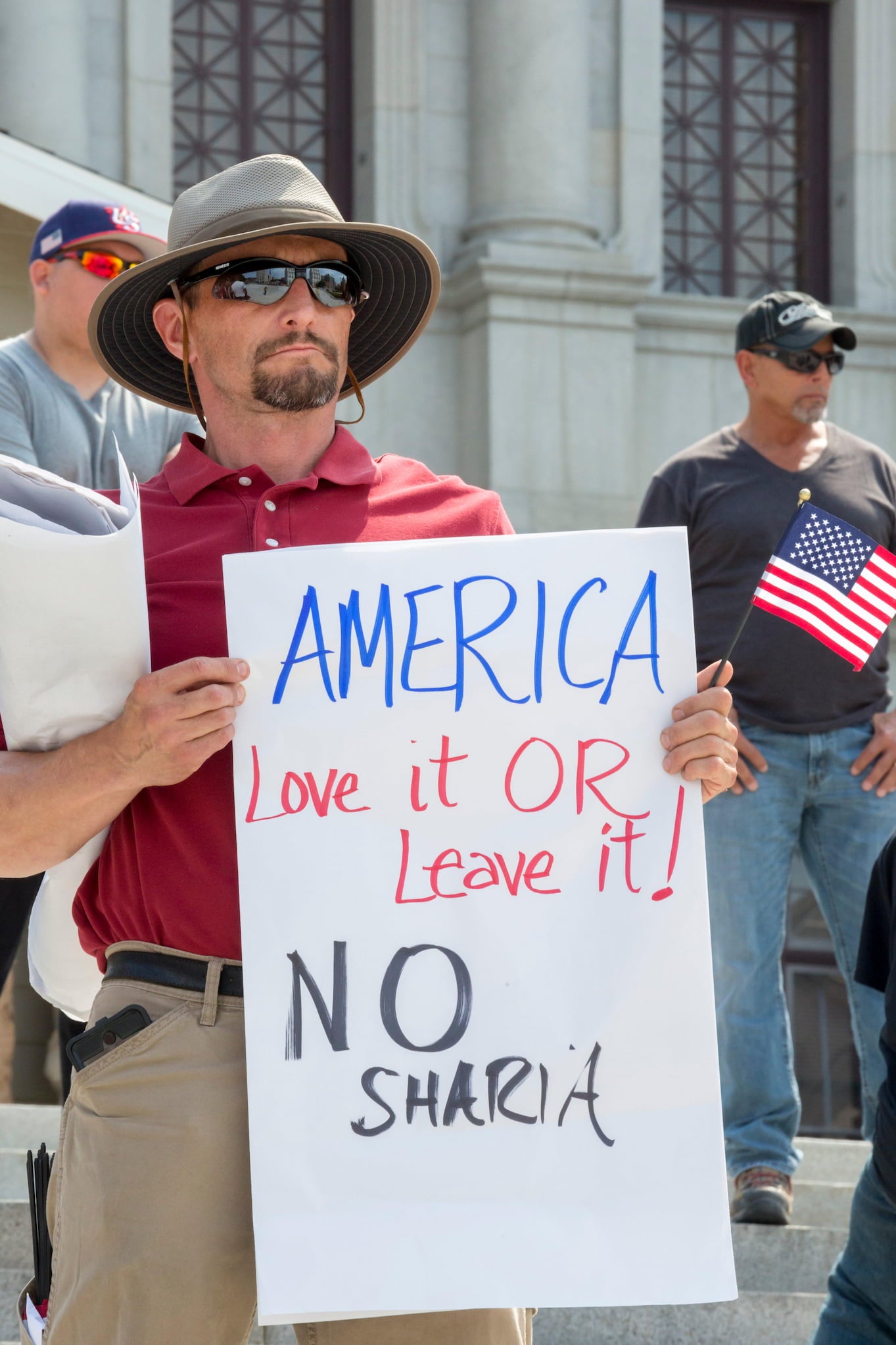 On the steps outside of the Pennsylvania State Capitol, a white man with a goatee holds a sign that reads, "America love it or leave it! No Sharia."