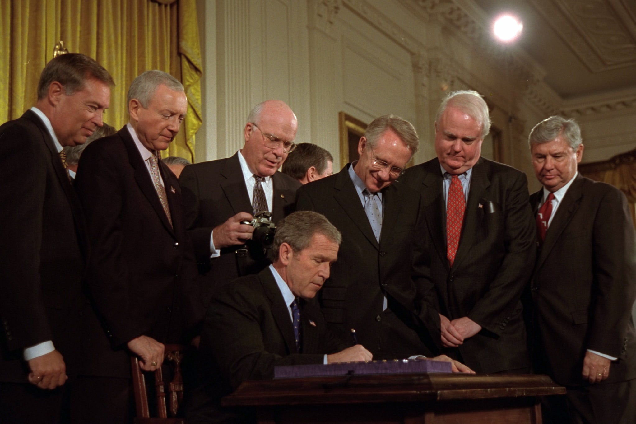 President George W. Bush, seated at desk, signs the Patriot Act into law. Six white men dressed in suits stand in half-circle around him.