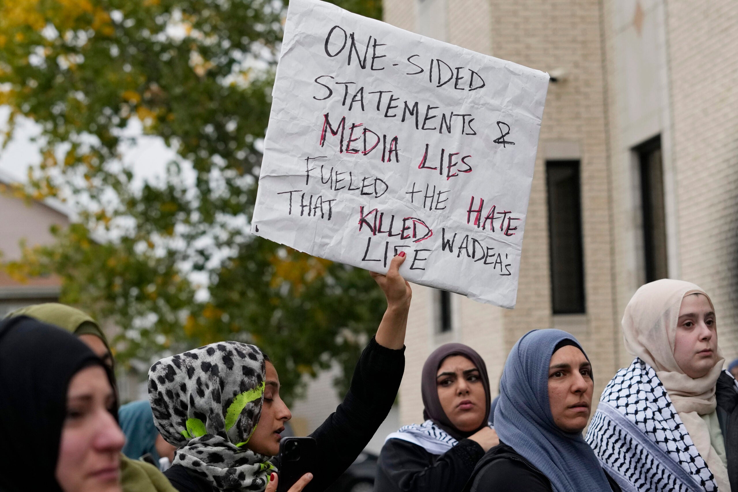 A Muslim American holds a sign that reads, "One sided statements and media lies fueled the hate that killed Wadea's life." Others stand with her.