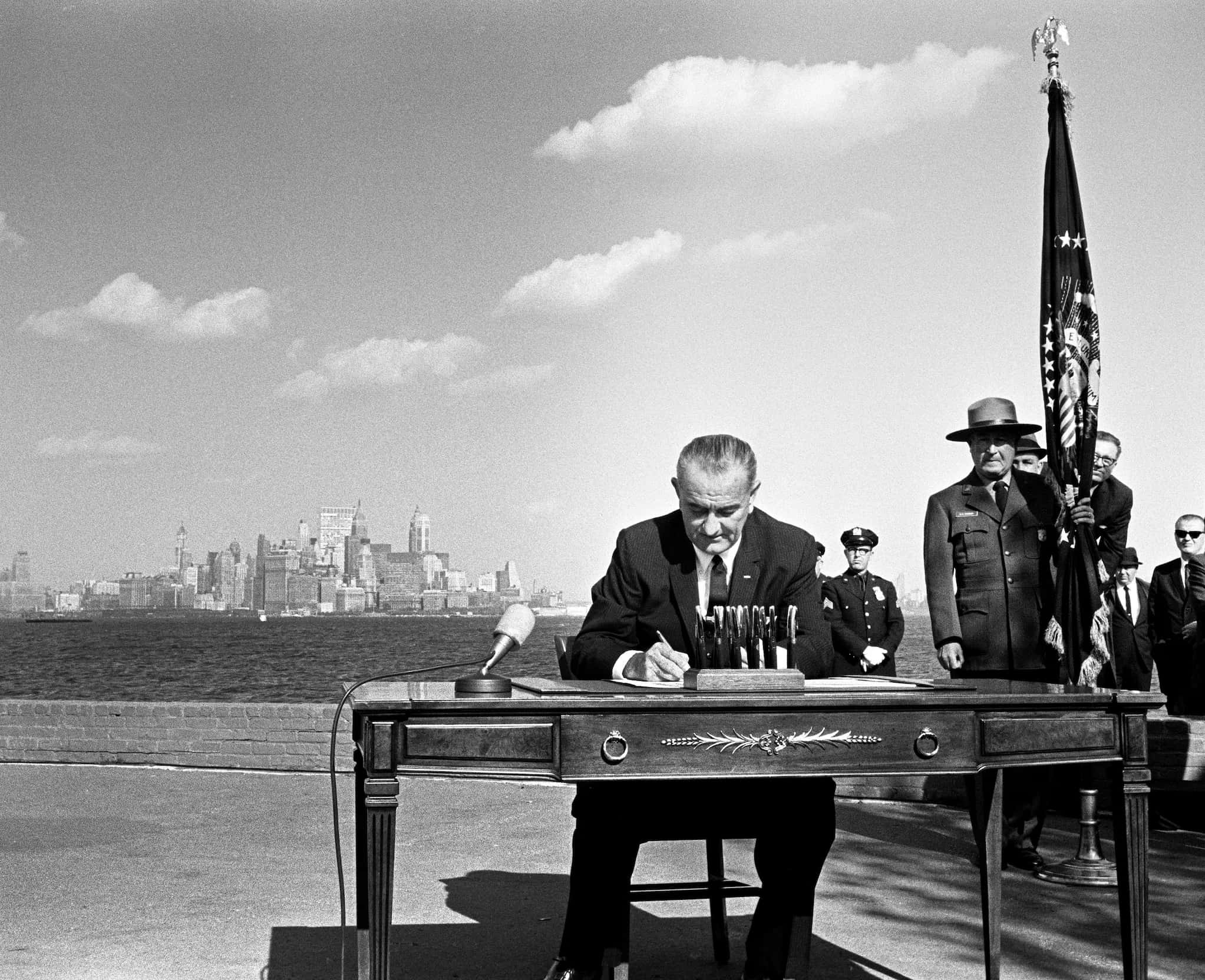 President Lyndon B. Johnson sits at a table on Liberty Island with New York City skyline behind him as he signs the Immigration and Nationality Act.