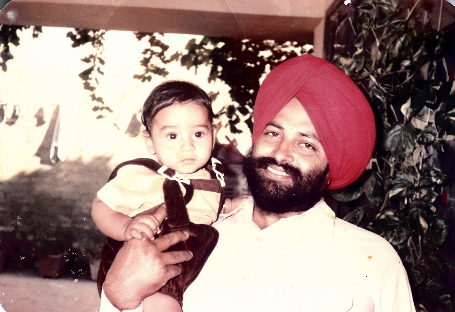A smiling and bearded Balbir Singh Sodhi, wearing a red dastār turban and white button down shirt, carries his toddler nephew in his right arm.
