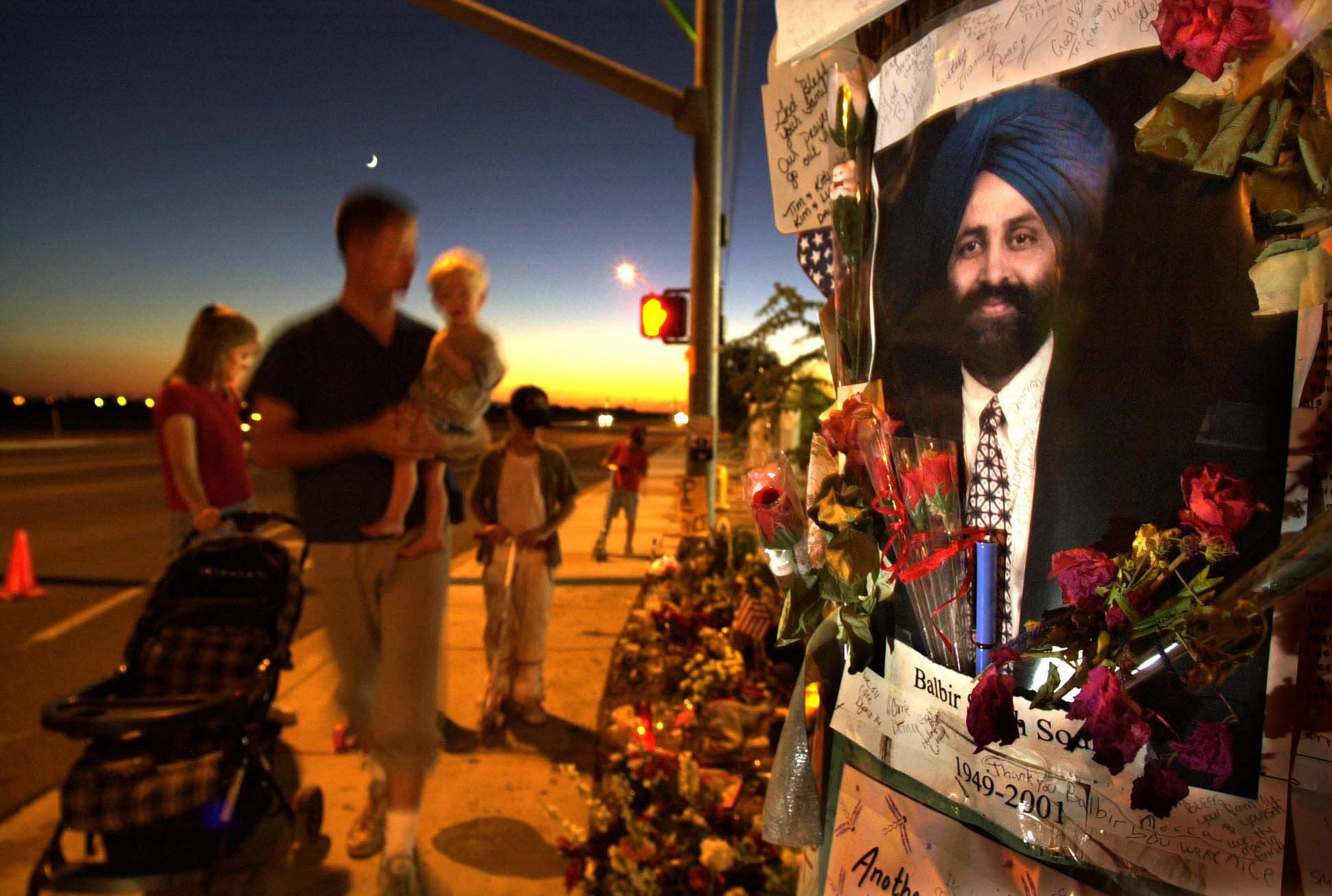 A family of five visits Balbir Singh Sodhi's memorial at dusk. Flowers, candles, photos, and notes surround his photograph set up by his gas station.