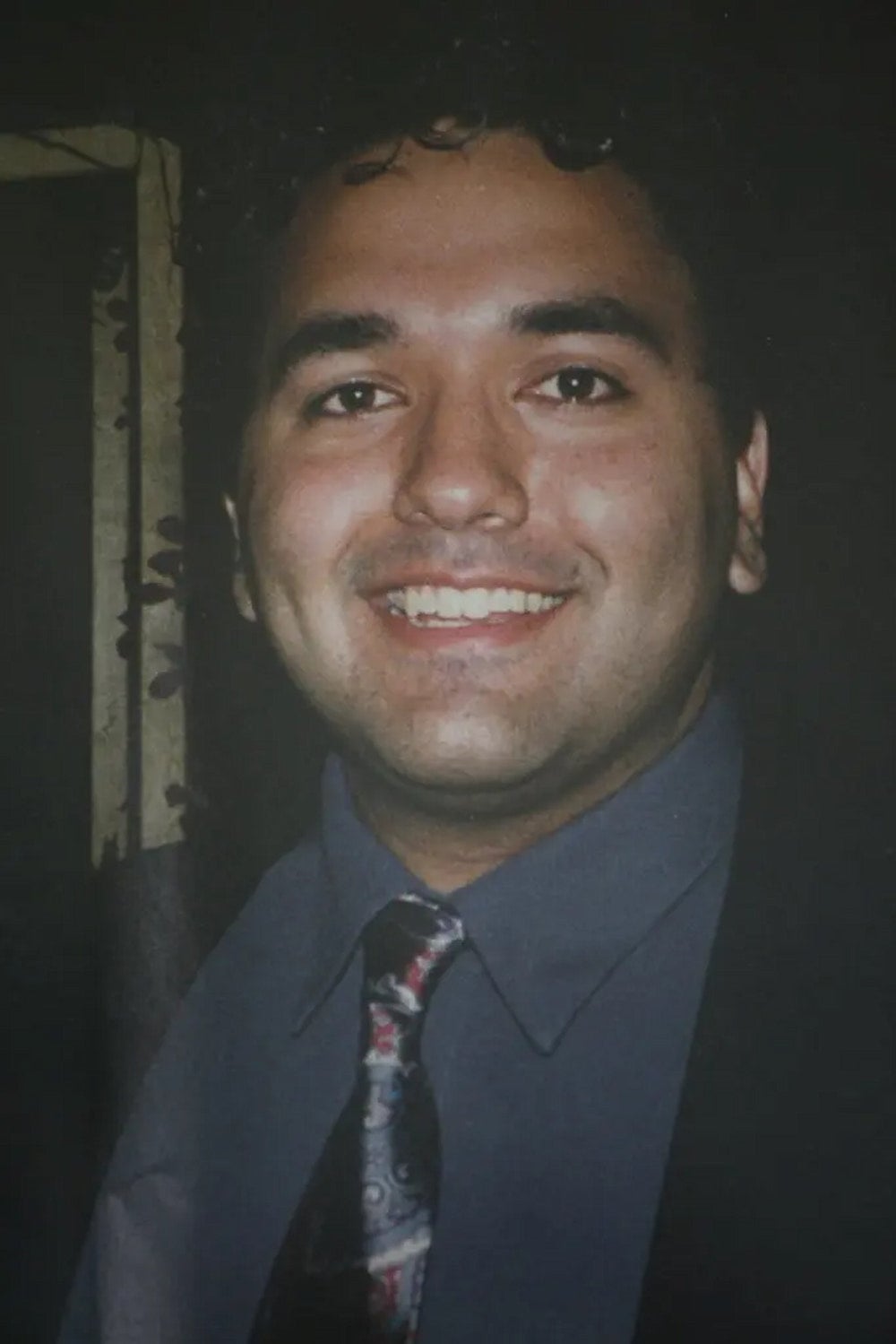 Mohammad Salman Hamdani, a Pakistani American man with curly hair wearing a dark suit with a paisley patterned silk tie, smiles at camera.