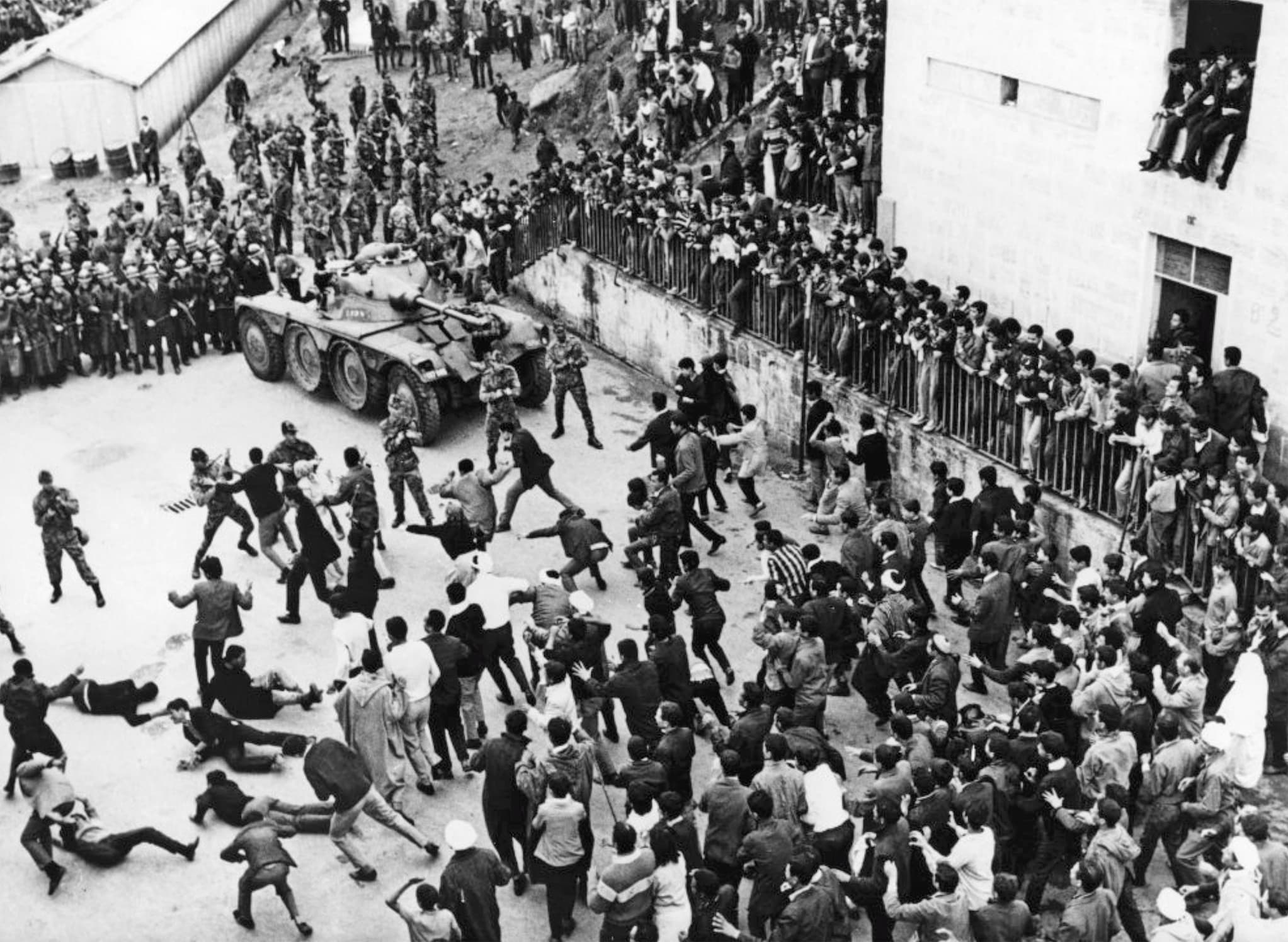 High angle still from The Battle of Algiers shows a fight between citizens and soldiers with a tank. Onlookers crowd around to watch the fight.