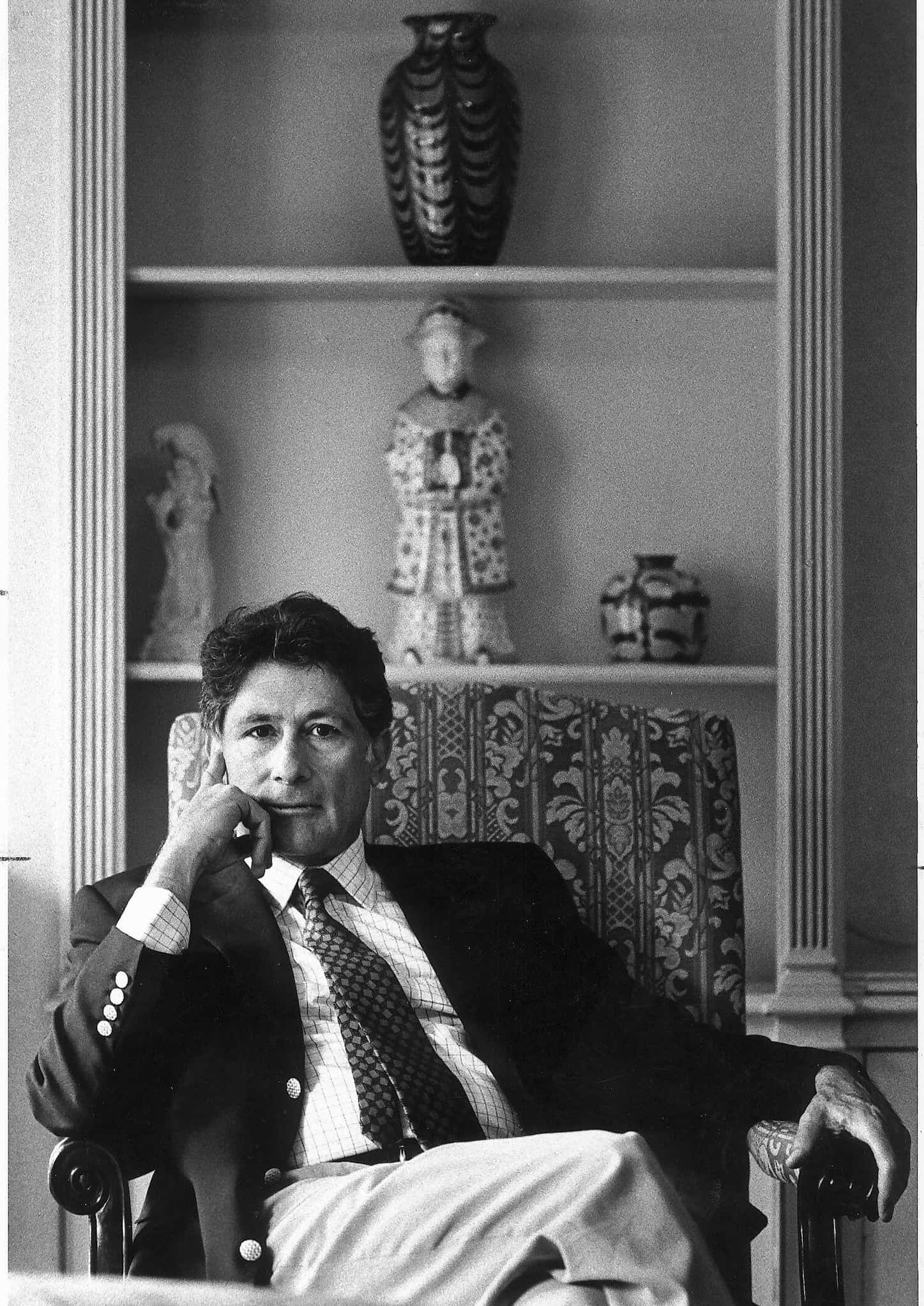 Edward Said, dressed in a suit, sits in an arm chair and rests his head on his hand. Behind him is a bookshelf displaying ceramic statues and vases.