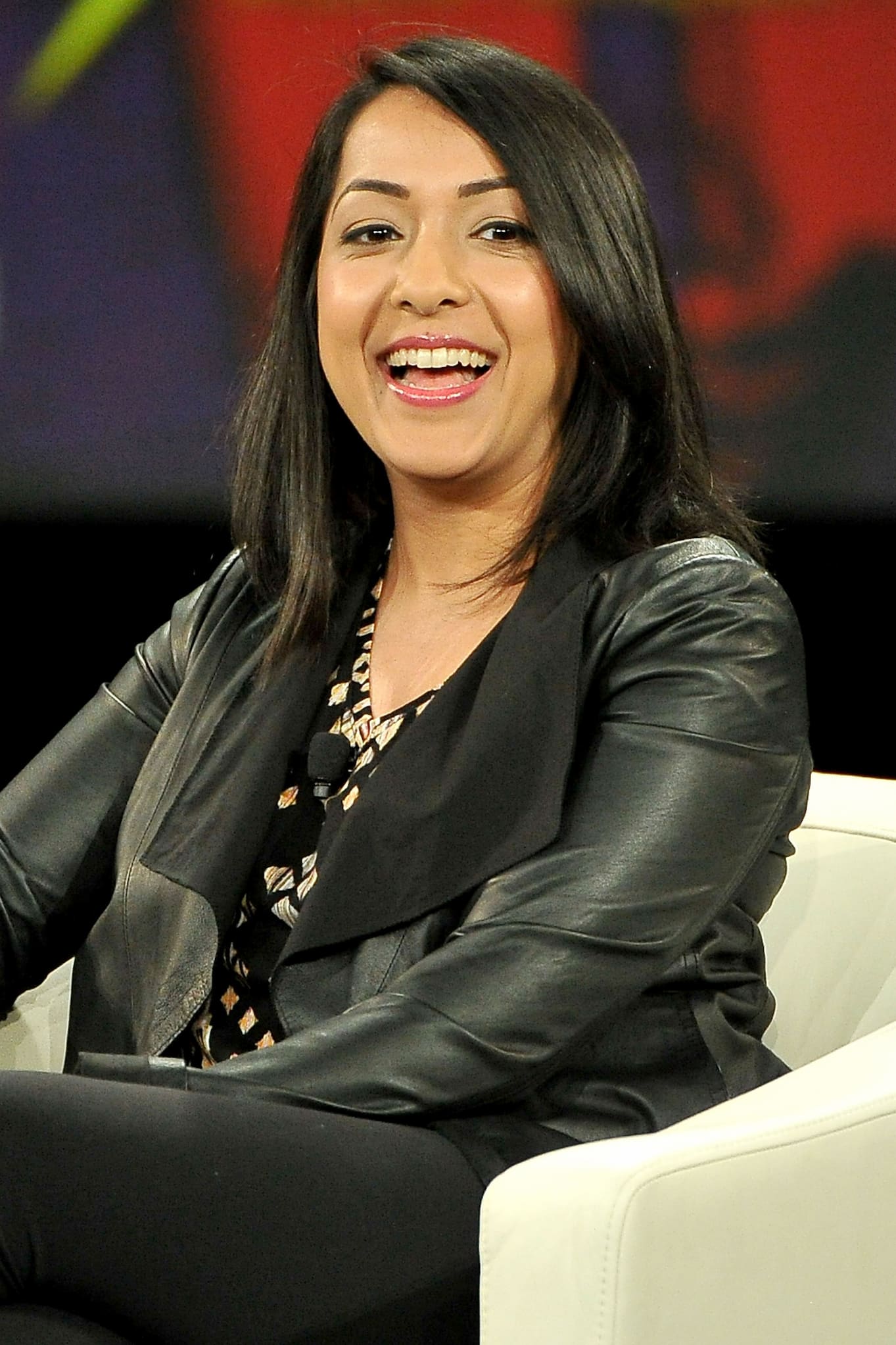 Sana Amanat, a Pakistani Muslim American, smiles while seated on stage. She wears a pleather jacket, a patterned blouse, and pants.