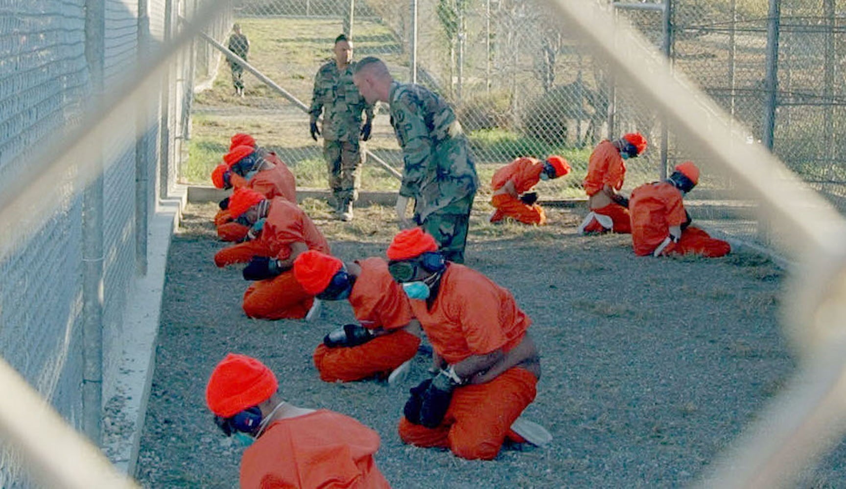 Ten prisoners, wearing jumpsuits, beanies, masks, handcuffs, earmuffs, and blindfolds, kneel as they are guarded by military police.