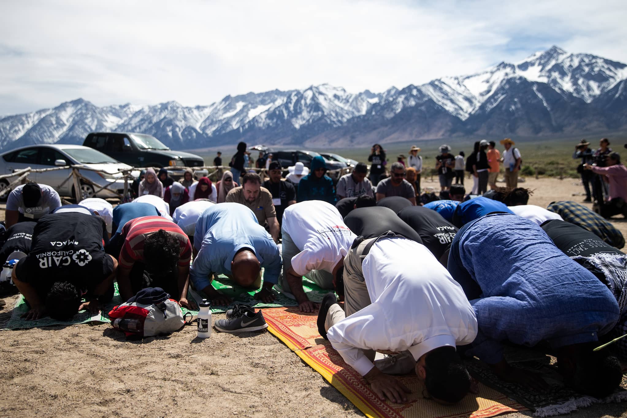 A group of Muslims kneel and bow on prayer mats during outdoor prayer. In the background are large, snow-capped mountains.