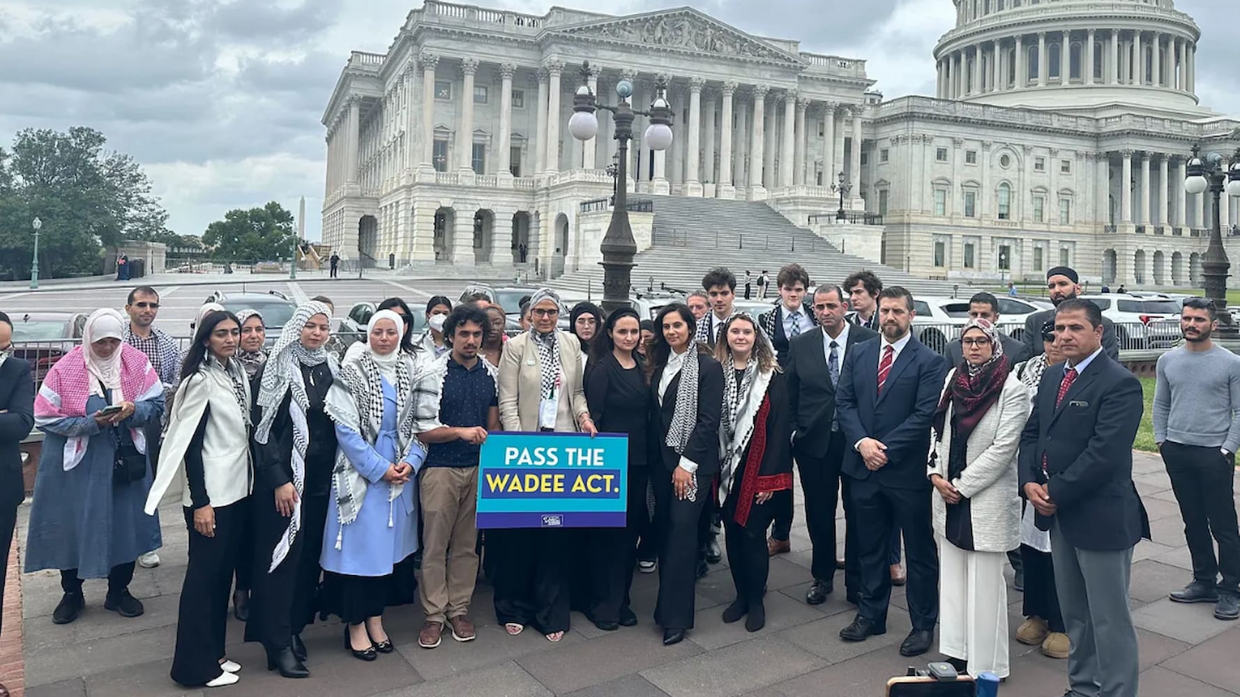 A group of men and women gather outside and stand behind a blue, purple, and yellow sign that reads, "Pass the Wadee Act."