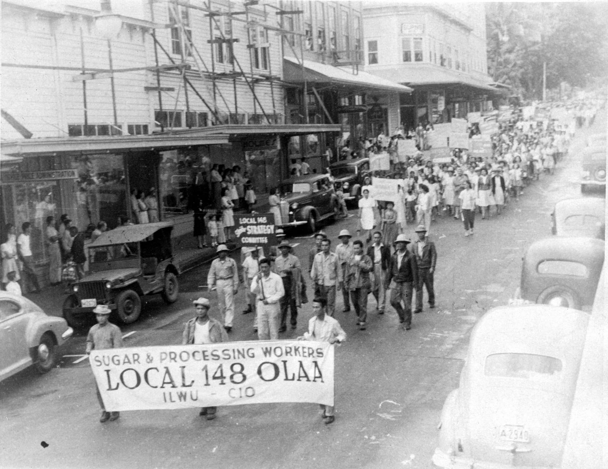 A protest led by Local 148 O L A A spans the length of a car-lined main street. Spectators on the sidewalk stand in the shade of scaffolding.