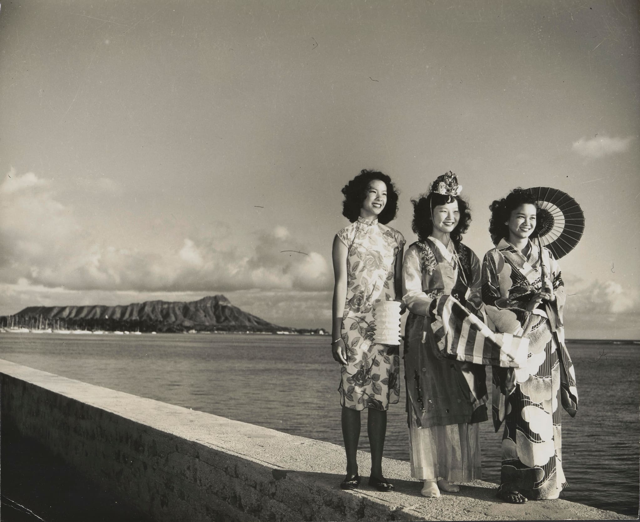 Three Asian women representing each of their cultures with traditional dress stand on a sea wall aside the ocean. Diamond Head is in the background.