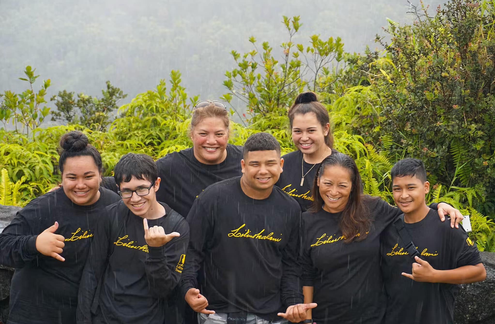 A group of smiling teens wearing matching long-sleeved black T-shirts pose in front of a forested mountain while throwing the Shaka sign.